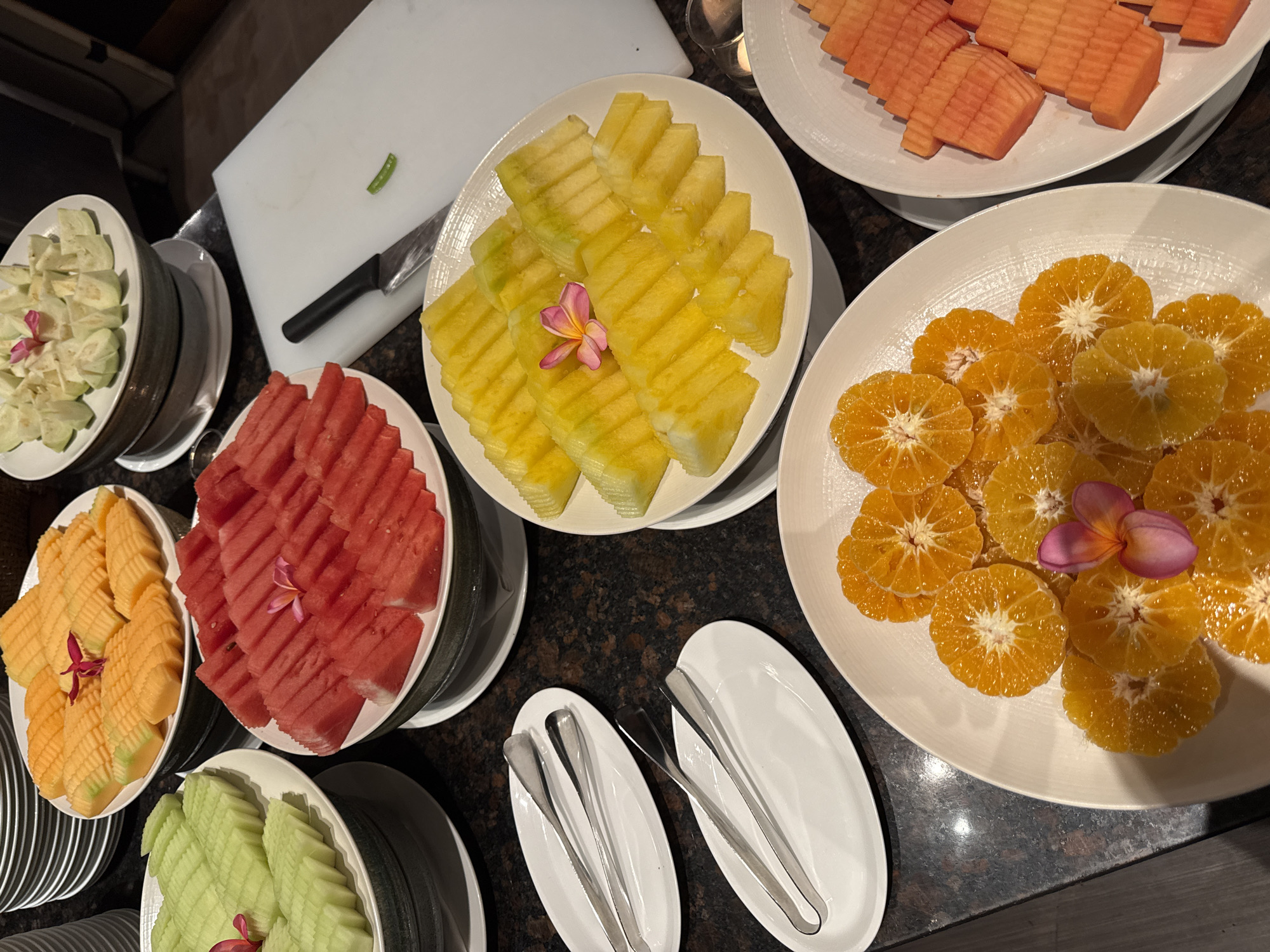 This is a top-down, slightly angled shot of a fruit buffet, likely in a restaurant or hotel in Kuta Selatan, Indonesia. The dark, speckled countertop is visible beneath several platters of sliced fruit, including what appears to be cantaloupe, honeydew melon, watermelon, pineapple, orange slices, and papaya. There are also a few decorative pink and orange flowers placed on some of the fruit platters. Two pairs of metal serving tongs rest on white oval plates to the left of the frame. In the background, a white cutting board with a knife and a small green chili pepper is visible, along with stacks of white plates and bowls. There are no people visible in the image.