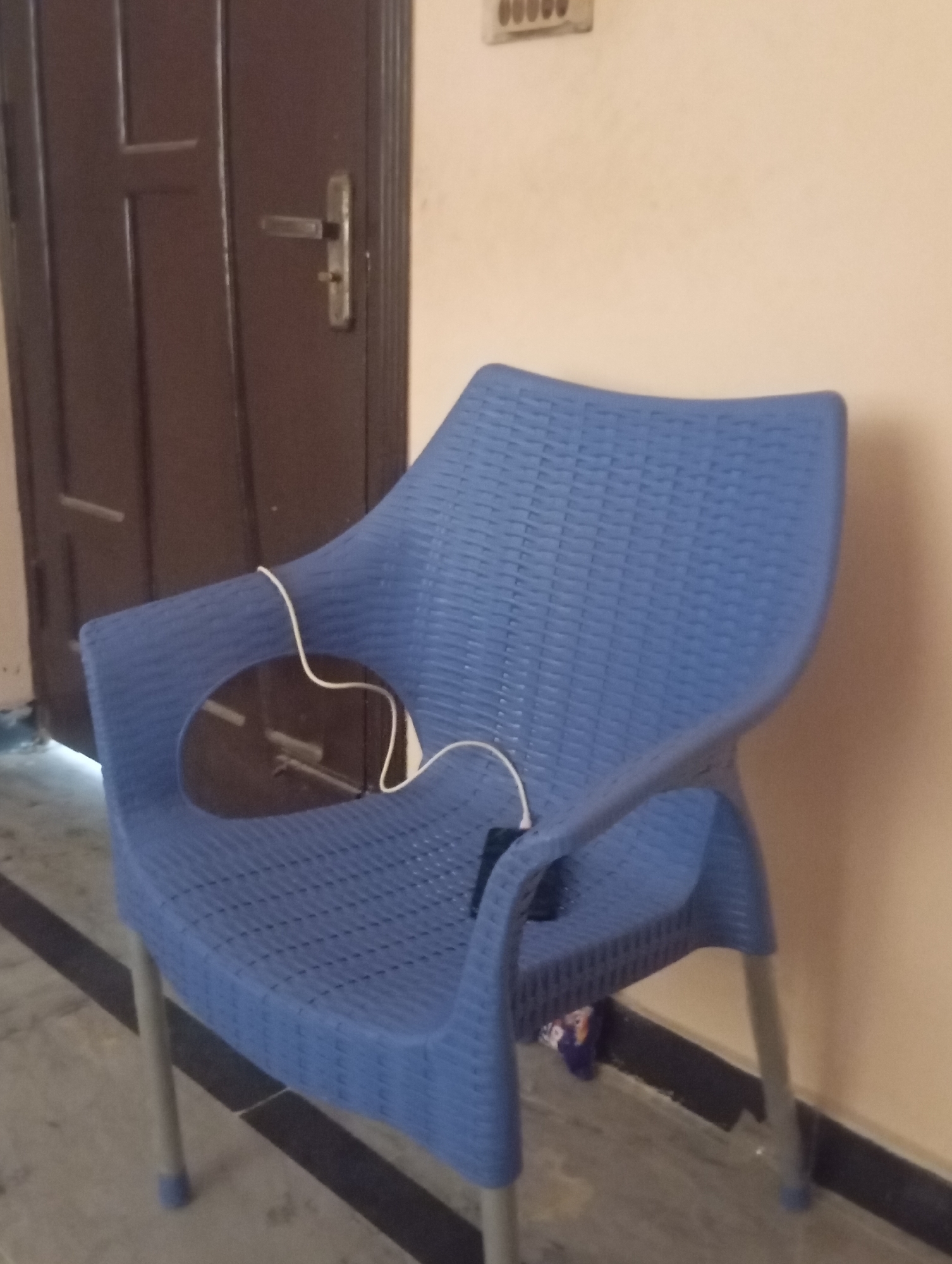 A blue plastic chair is positioned in front of a brown wooden door, with a beige wall to the right. A white wire is draped over the armrest of the chair. The floor appears to be tiled with a black border. A ventilation grate is visible on the wall above the door.