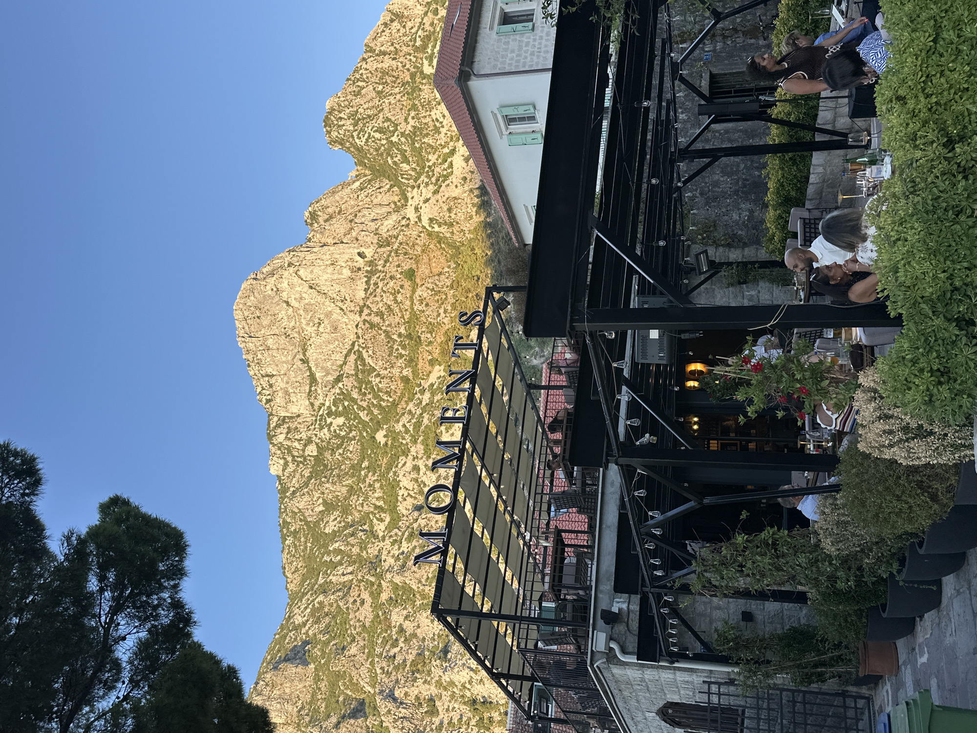 The photograph depicts an outdoor dining area of a restaurant named "MOMENTS" in Dobrota, Montenegro. The restaurant features a multi-level structure with a covered patio. The upper level has tables and chairs arranged under a canopy, while the lower level also has outdoor seating. The restaurant's name is prominently displayed in large, dark letters on the upper level's facade.
Several patrons are seated at tables, engaged in conversation and dining. Some appear to be couples, while others are in small groups. A woman in a brown top is standing and speaking to someone seated at a table. The background is dominated by a rugged, mountainous landscape, with the peaks illuminated by the warm light of the setting sun. A white building with green shutters is visible to the right of the restaurant. Lush green plants and trees are strategically placed around the dining area, adding to the ambiance. The sky is clear and blue.