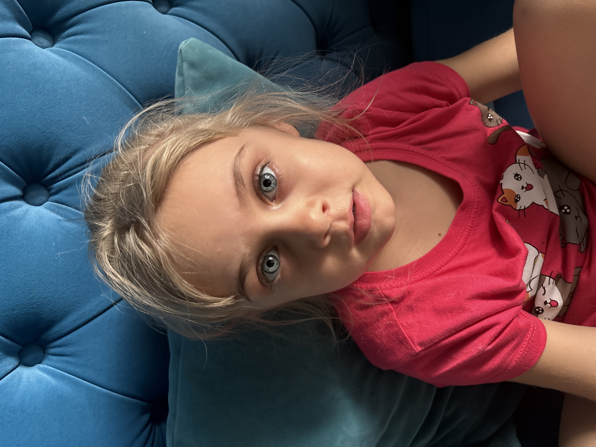 A young girl with fair skin, blonde hair, and striking blue eyes is lying on a blue tufted cushion, possibly a couch or lounge. She is wearing a red t-shirt with cartoon cat designs. The background and setting suggest an indoor environment, possibly a home.