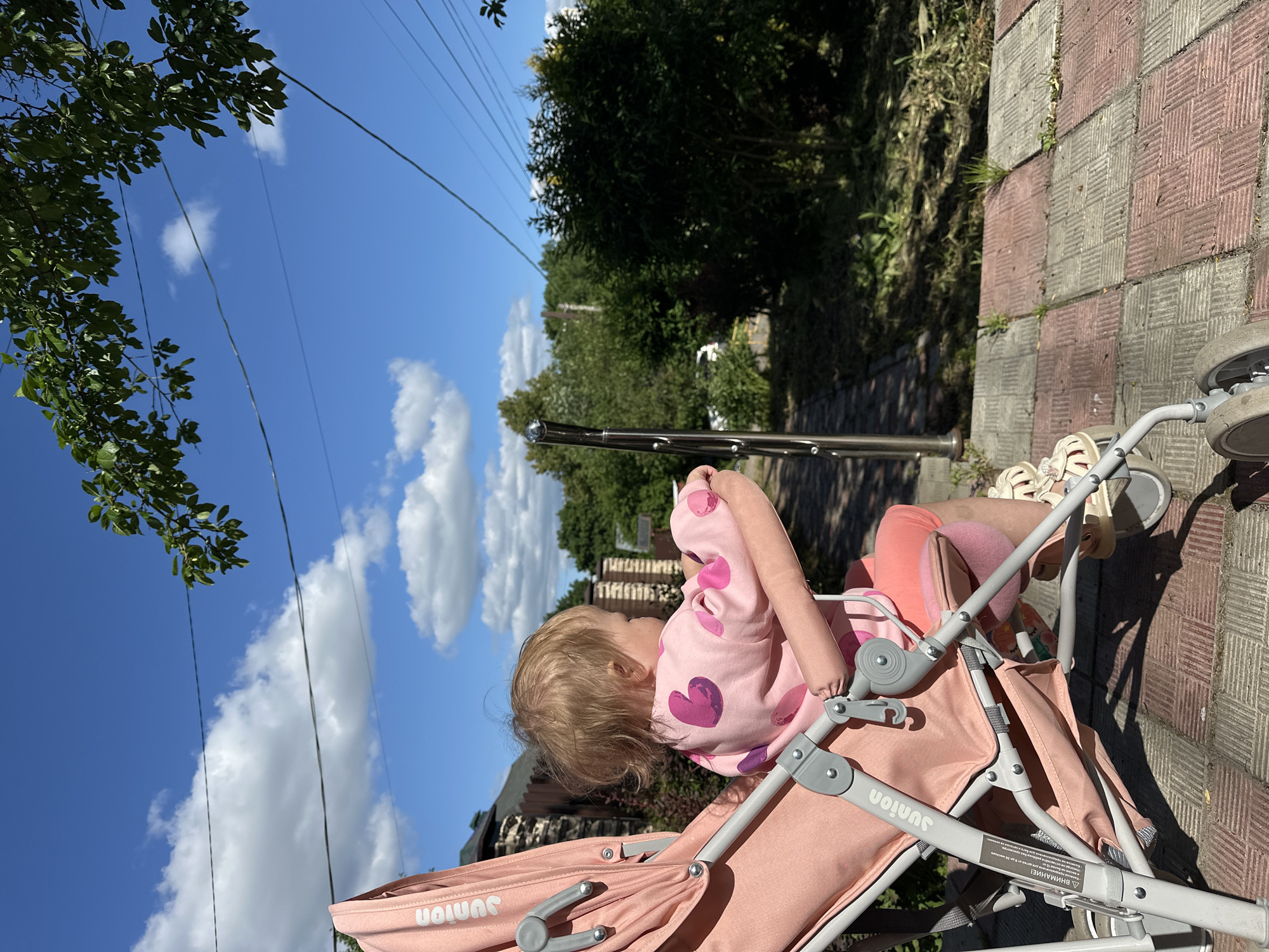The image depicts a young child seated in a pink stroller on a bright, sunny day. The scene is captured outdoors, likely on a paved path or sidewalk with a distinctive checkered pattern of alternating grey and reddish-brown square tiles.
The child, appearing to be a toddler with fair hair, is dressed in a pink long-sleeved top decorated with darker pink heart patterns, light pink bottoms, and white sandals. They are positioned in the stroller, facing generally forward, but their head is turned to the right, observing something in the distance.
The pink stroller features the brand name "Junior" clearly visible on its fabric, and a white warning label on the frame reads "ВНИМАНИЕ!" (Attention!). In the background, lush green trees and foliage are abundant, suggesting a park-like or residential setting. Above, a vivid blue sky is dotted with scattered white cumulus clouds, and several thin electrical wires stretch across the frame. A metallic pole or railing stands upright in the mid-ground. The strong, clear lighting and distinct shadows indicate it is daytime. The overall impression is a peaceful moment, possibly during a casual outing or walk in Nizhny Novgorod, Russia.