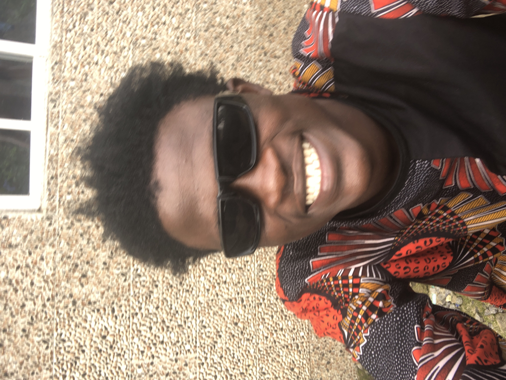 The image shows a close-up, head-and-shoulders portrait of a young Black male with dark, curly hair, wearing black sunglasses and a brightly colored shirt with a pattern of red, yellow, and black. He is smiling, revealing his teeth. The background appears to be a textured wall with small, multi-colored pebbles, and a portion of a window frame with white mullions is visible at the top. The setting is likely outdoors, possibly in the city of Ado, Nigeria, given the style of the wall and the natural lighting. The overall impression is one of casual portraiture.