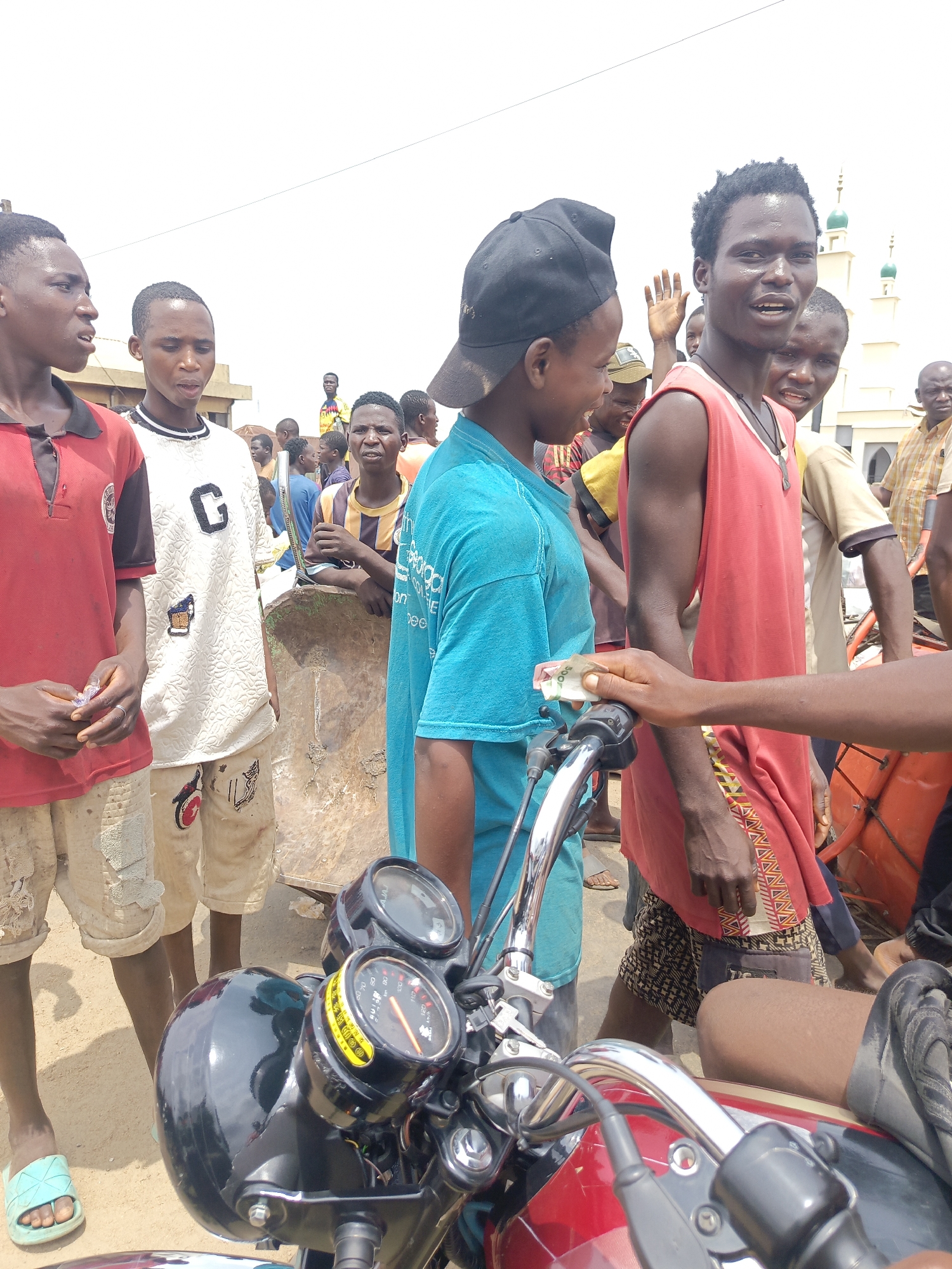 A street scene in Akwanga, Nigeria depicts a group of young men gathered around a red motorcycle. Several of the men are wearing casual clothing, including t-shirts and shorts. One man in a blue shirt is wearing a backwards baseball cap. A man in a red tank top is facing the camera and smiling, while holding up his hand. A hand holding a folded Nigerian Naira note is visible near the motorcycle handlebars. A mosque with green domes is visible in the background. The overall scene appears to be a casual gathering in a public space.