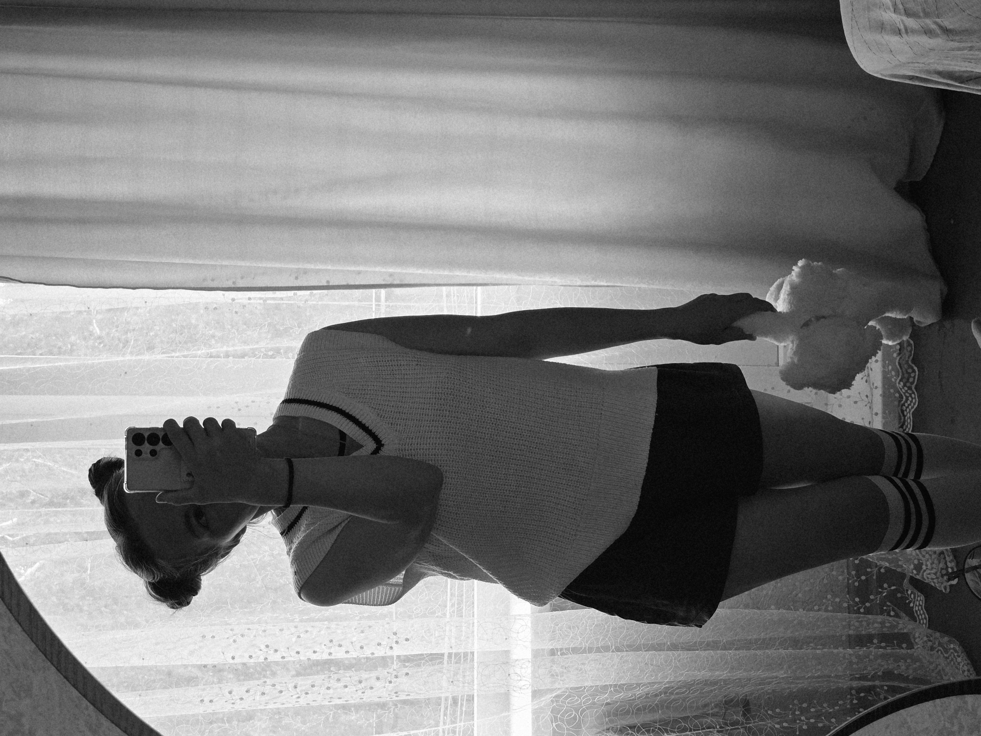 A black and white, full-body indoor image depicts a person standing and holding a smartphone, capturing a mirror selfie. The person's face is largely obscured by the smartphone, held in their left hand, revealing one visible eye. Their dark hair is styled in two distinct buns on top of the head. They are wearing a light-colored sleeveless V-neck knit vest with dark horizontal stripes on the collar, dark shorts, and light-colored knee-high socks with three dark horizontal stripes. A dark band or bracelet is present on their right wrist. The person's right hand holds a light-colored, fluffy, possibly plush toy, which dangles downwards. The background features a window covered by sheer, patterned white curtains on the left, displaying floral or leafy motifs, and a plain, light-colored drape pulled across on the right. A dark floor is visible, and the curved edge of a mirror frame is present on the far left. A piece of furniture with draped fabric is partially visible on the bottom right. The scene is backlit by the window, creating a partial silhouette of the subject.