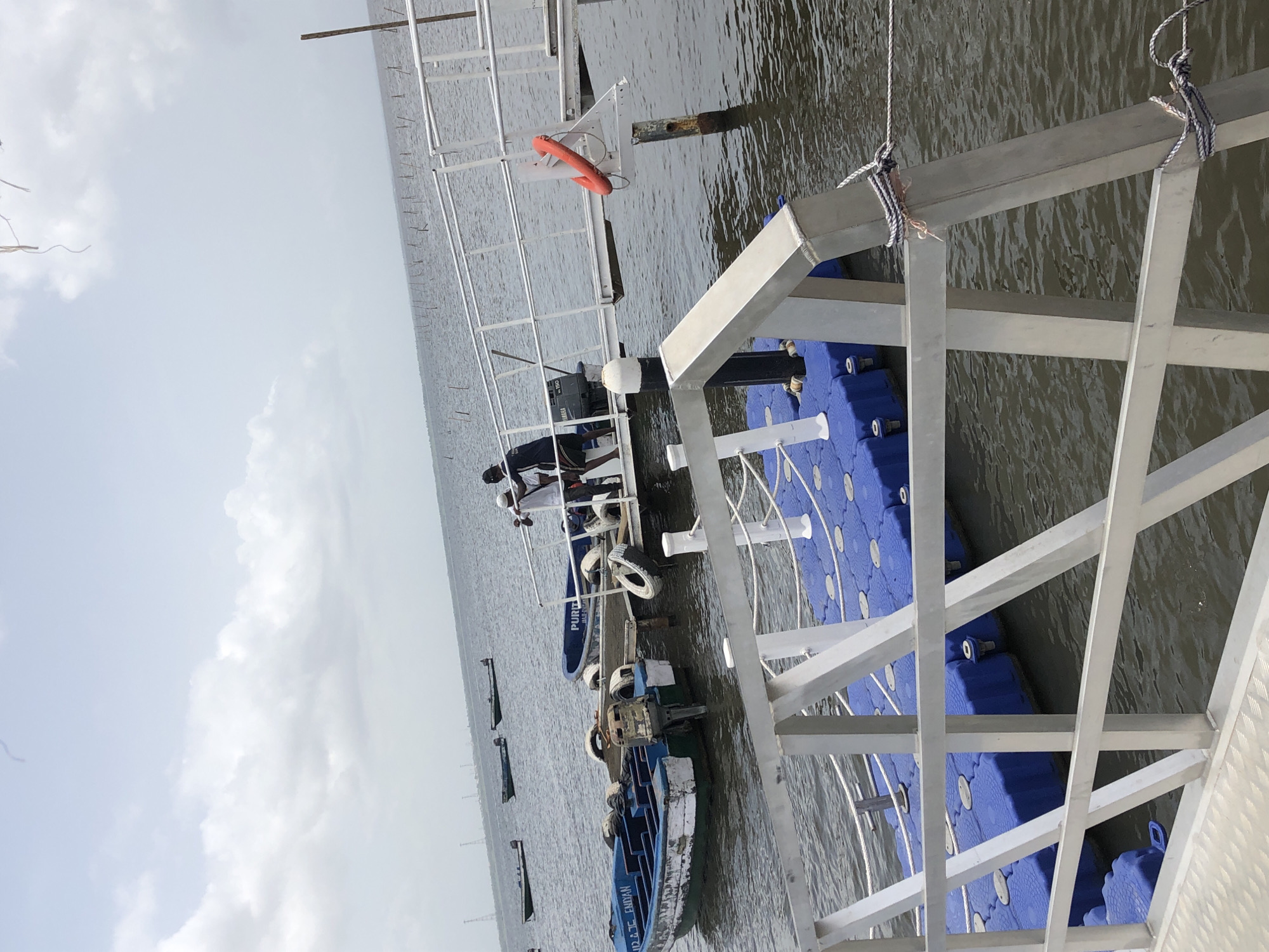 The image depicts a waterside scene, likely a dock or pier area on a calm body of water, possibly a bay or estuary, under an overcast sky.
In the foreground, a modern modular floating dock is visible, constructed from blue interlocking plastic pontoons and supported by a white or silver-colored metal framework. Ropes are tied to various parts of the metal structure, some extending downwards towards the water or other elements. An orange lifebuoy hangs from a part of the metal framework.
Several small boats are moored near the dock. One boat, painted blue and white, has visible text "PURBA CHANDAN" on its side. Another blue boat further down the dock has "PURBA" and "JAGERIA" (partially visible) written on it and is equipped with an outboard motor, identifiable by the "YAMAHA" brand and "150" (possibly indicating horsepower) text. Several old tires are used as fenders on the boats.
At least two people are visible in the scene. One person, wearing a dark shirt and light pants, is standing on the blue and white boat near the dock. Another person can be seen further along the dock structure. Their activity appears to be related to the boats or maintaining the dock, though specific actions are not clearly discernible.
The water, reflecting the overcast sky, shows ripples on its surface. In the background, numerous thin poles or stakes extend into the distance across the water, suggesting aquaculture, fishing nets, or markers. The horizon line between the water and the sky is flat and expansive. The sky is largely covered with light gray clouds, indicating an overcast or partly cloudy daytime setting. The overall impression is one of a working waterfront.