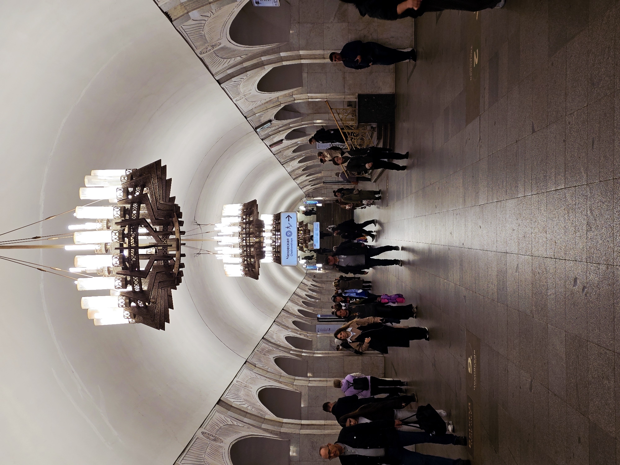 Captured at Chekhovskaya metro station in Moscow, Russia, the image showcases an underground platform bustling with people. Overhead, ornate chandeliers illuminate the vaulted ceiling with its decorated arches. Individuals of varied ages and appearances are seen traversing the platform, some standing and waiting, while others are walking. The floor is tiled, reflecting the ambient light. Signage is visible, indicating directions within the station.