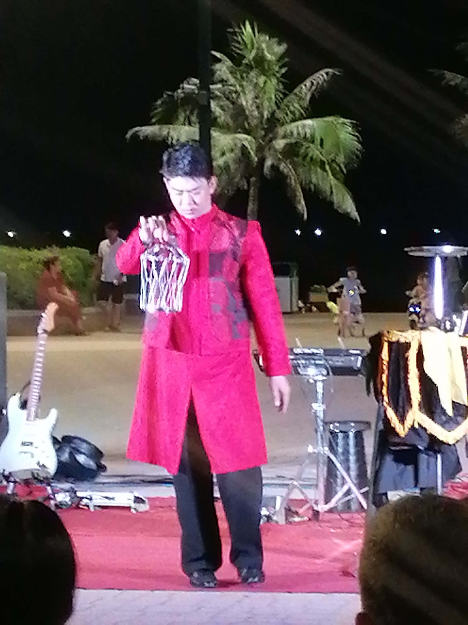 The image shows an Asian man on a stage outdoors at night in Nha Trang Ward, Vietnam. The man is wearing a red long-sleeved jacket over black pants. He is holding a white wire cage-like object in his left hand. In the background, there are palm trees. There are other people on the beach in the background, including children playing. A white electric guitar is on a stand near the left edge of the stage, and a keyboard stand and other equipment are also visible. The stage is covered in a red carpet. Part of the back of the heads of two people in the audience are visible at the bottom of the image.