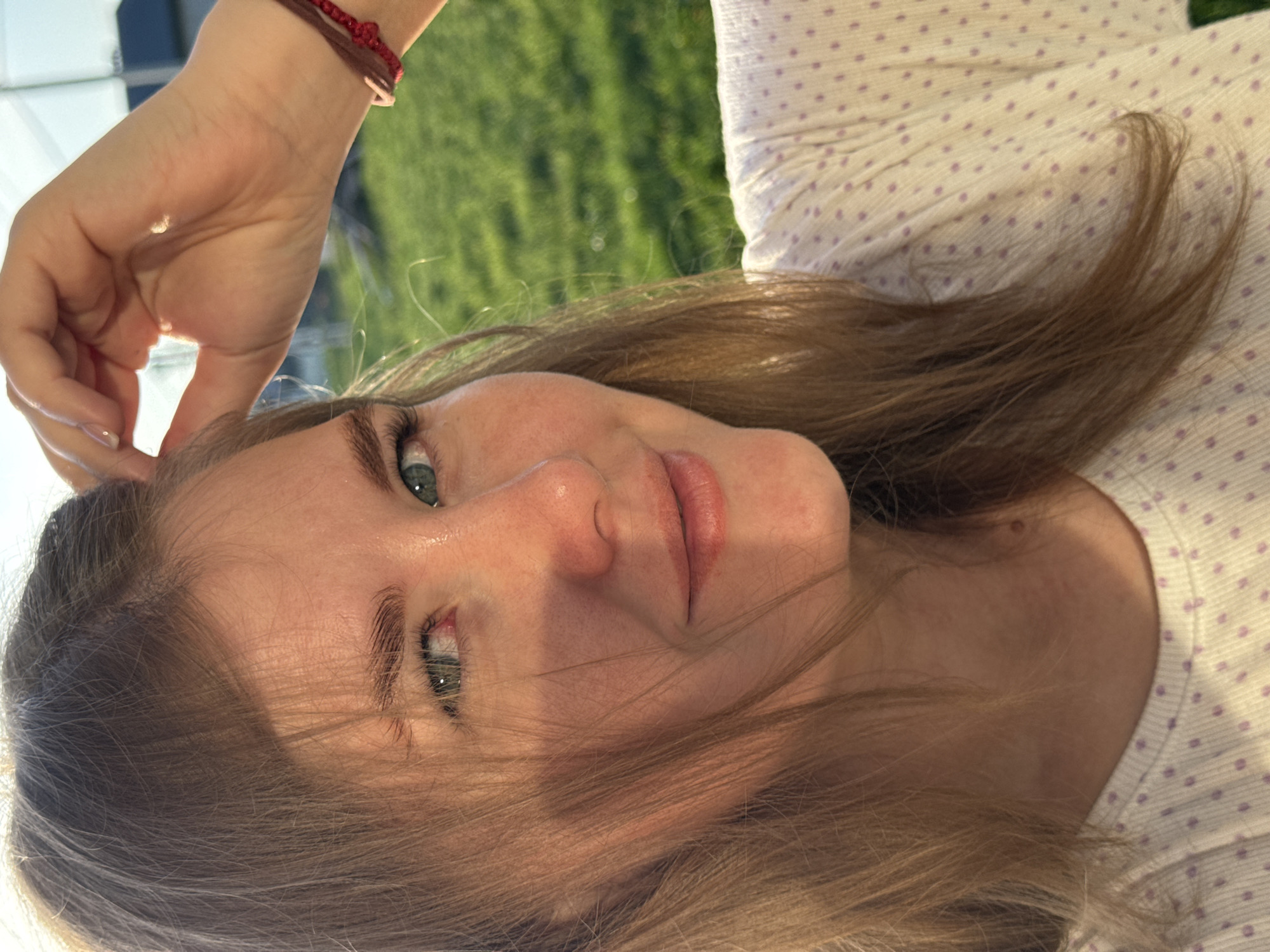 A young, fair-skinned woman with light brown hair and green eyes looks at the camera with a slight smile. She is wearing a white shirt with small lavender dots. Her left hand is raised near her forehead and she is wearing a bracelet with brown and red strings. The background shows a green, grassy area, possibly suggesting an outdoor setting near Ферма, Russia, although this is just a possible location based on the user's request.