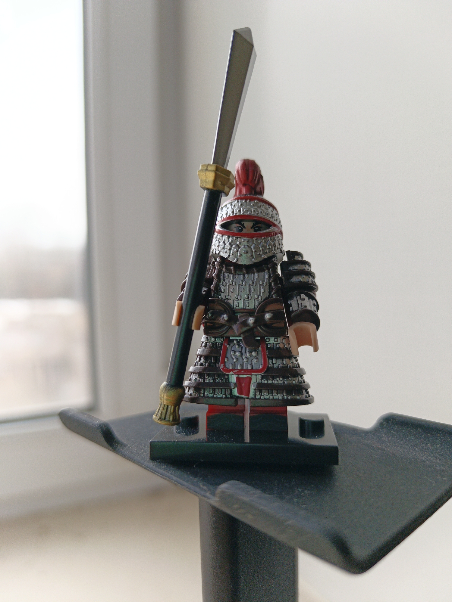 A close-up shot depicts a Lego minifigure adorned in highly detailed, Chinese-inspired armor. The figure wields a tall glaive-like weapon, its blade a shiny metallic color and the staff featuring golden accents. The armor is intricate, showcasing many plates and red underlinings and accents. The minifigure stands on a dark grey, possibly plastic, stand that is raised above the surface below. The background reveals a blurred view of a window, with light seeping through and a glimpse of the outside cityscape of Unknown, Unknown. The overall lighting of the scene is soft, illuminating the details of the minifigure.