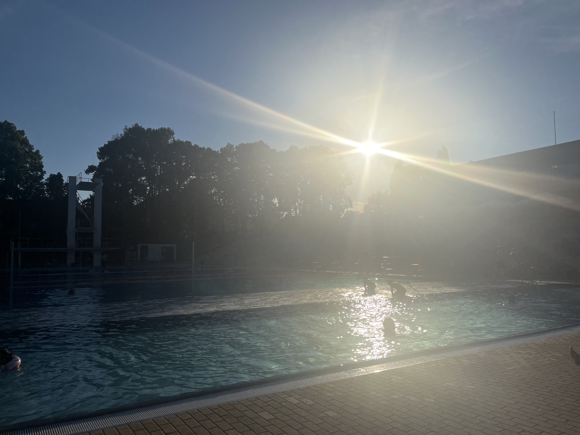 The image depicts an outdoor swimming pool bathed in bright sunlight, causing lens flare that obscures parts of the scene. In the foreground, a tiled poolside transitions to the clear blue water of the pool. Several people are visible in the water, some swimming or playing. In the background, trees form a dark silhouette against the sky, and a diving platform is visible to the left. To the right, a building with a sloped roof is partially obscured by the glare. The sky is clear and blue, with the sun positioned high and bright, creating strong highlights on the water's surface.
