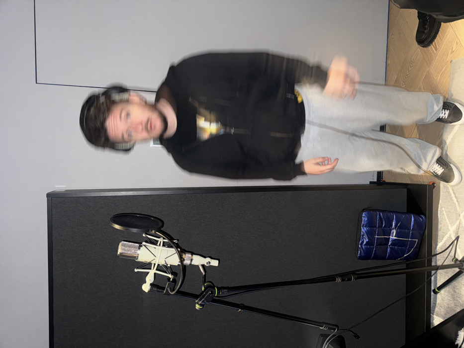 The image depicts an indoor recording studio or sound-treated room. A male individual, blurred due to motion, stands centrally. He wears black over-ear headphones, a dark zip-up hoodie over a dark graphic t-shirt, light grey sweatpants, and dark sneakers with white soles. His face is indistinct, but he appears to have short facial hair. To his left, a silver condenser microphone is mounted on a shock mount with a black pop filter, all supported by a black tripod stand. Behind the microphone is a tall, rectangular dark grey or black acoustic panel. A small, quilted blue bag rests on the floor next to the acoustic panel. The floor consists of light-colored wood or laminate on the right and a light-colored rug on the left. The walls are plain light grey. A partial view of a dark shoe and pant leg, belonging to another unseen individual, is visible in the bottom right corner. A rectangular white outline, possibly a doorframe, is visible on the far right wall. The scene is consistent with a recording session setup in the city of Khimki, Russia.