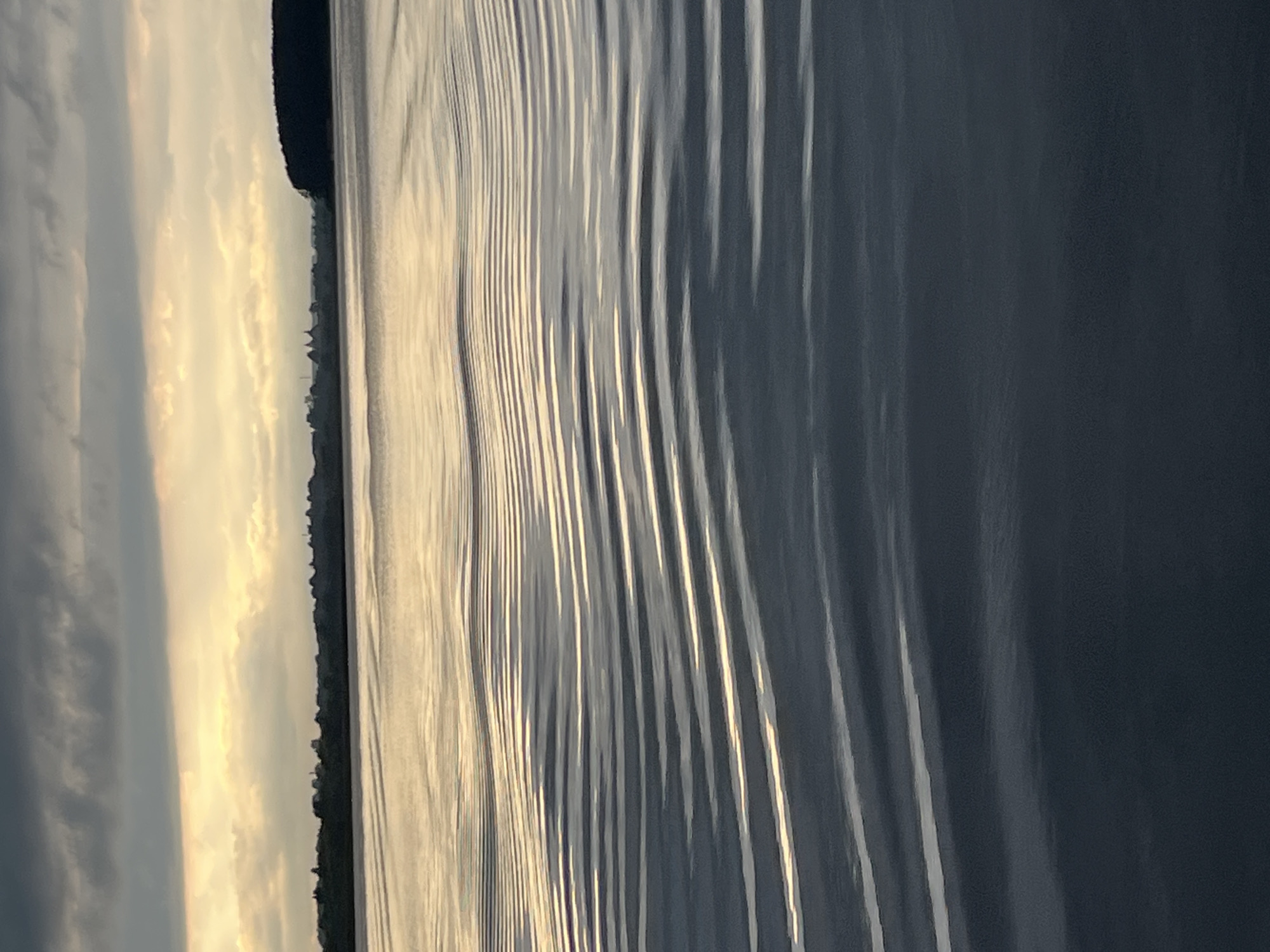 The image depicts a body of water with a shoreline visible in the distance. The sky is overcast with clouds, and the sun appears to be setting or rising, casting a warm glow on the water and clouds. The water is rippled, creating a pattern of light and shadow. The shoreline is lined with trees and some buildings. The location is identified as Radchenko, Russia. There are no people or active interactions visible in the image. The scene is serene and natural.