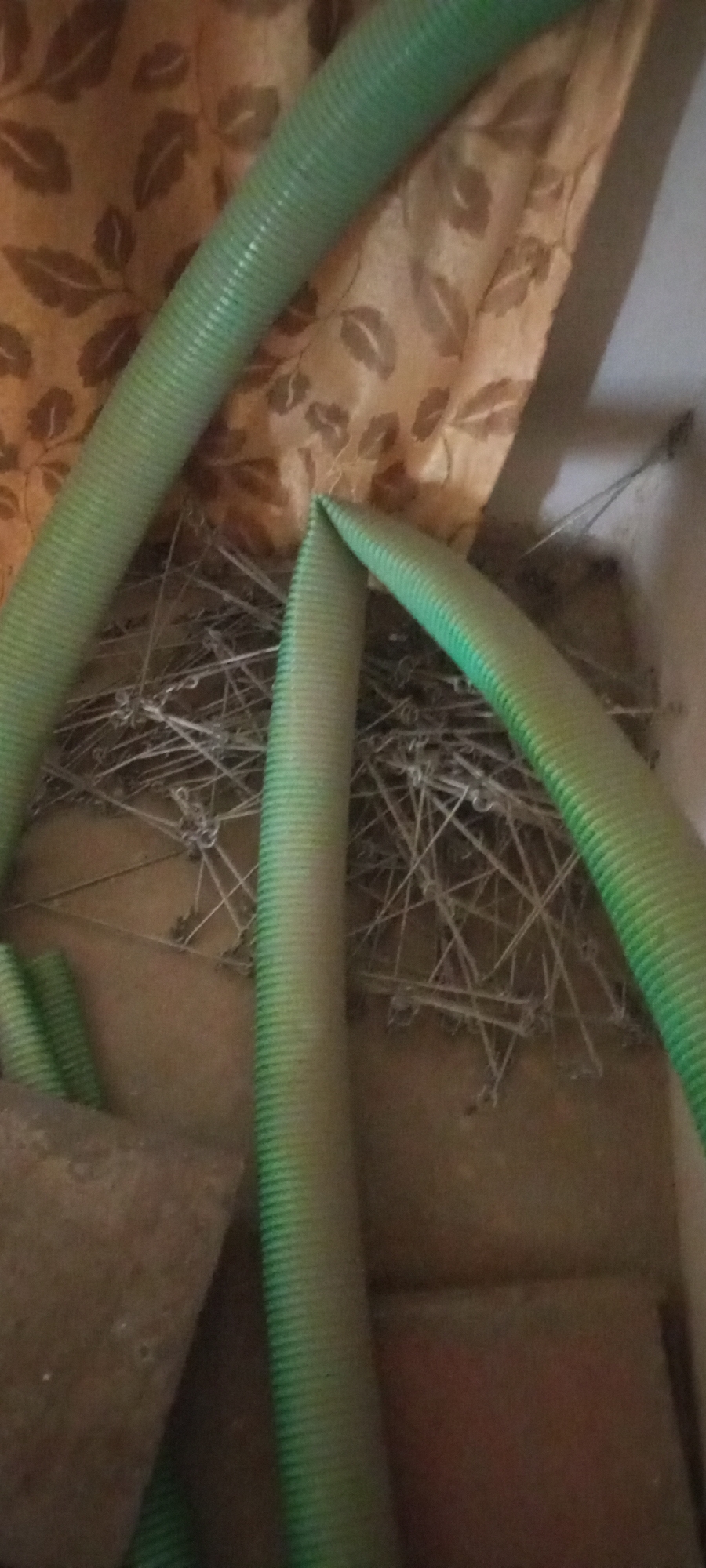 The image shows a close-up view of several green, ribbed hoses. In the background, a brown curtain with a leaf pattern is visible. The hoses are coiled and resting on a surface composed of numerous thin, metallic wires or staples, which appear to be entangled. There is also a brown, textured surface, possibly wood or cardboard, in the foreground. The scene is indoors, and the lighting suggests it might be a storage area or a workspace. The location is Jalingo, Nigeria.