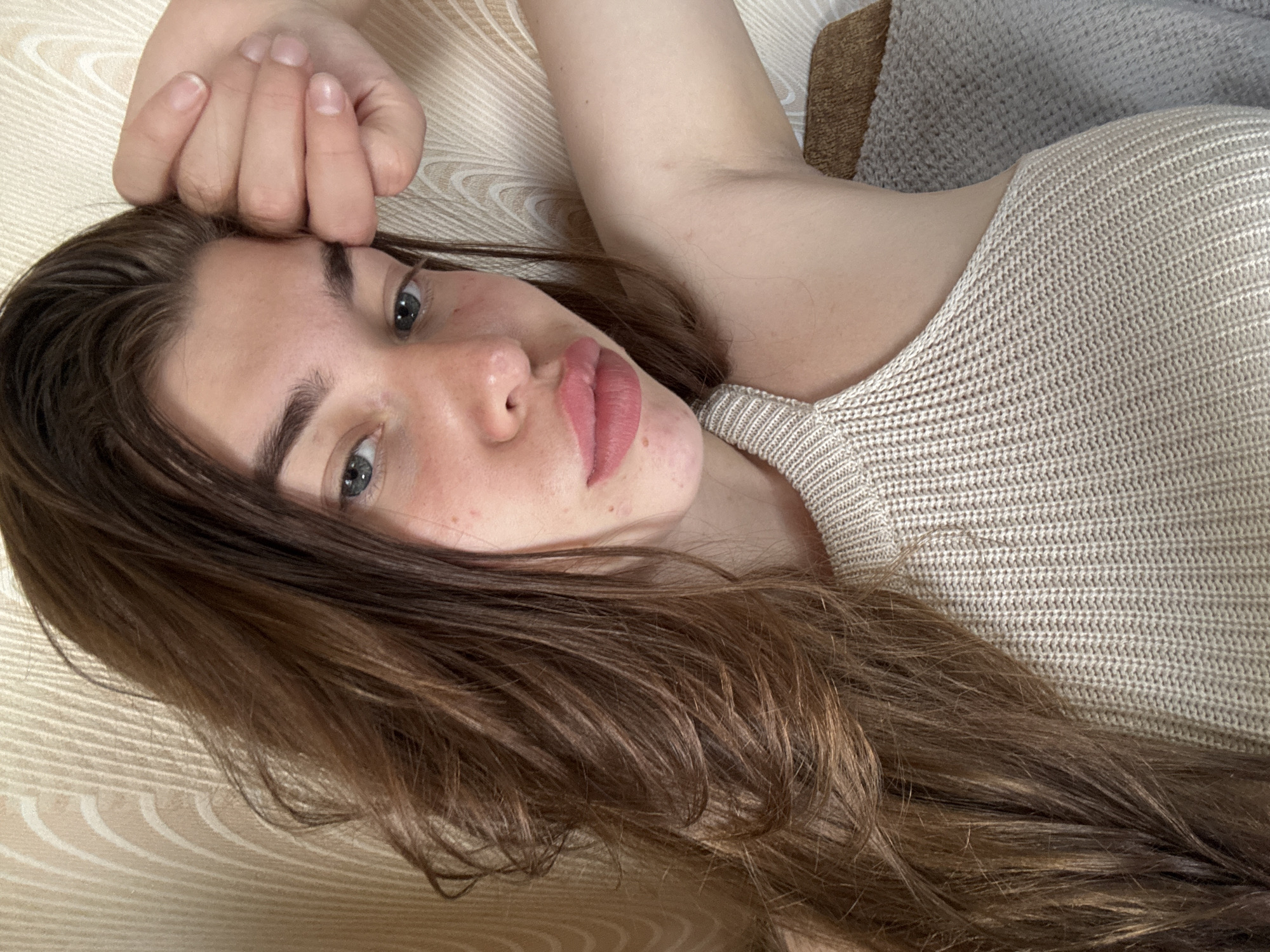 A young woman with long, brown hair and fair skin is captured in a close-up, selfie-style shot. She is wearing a beige, knitted top with a halter neckline. Her left hand is resting on her forehead, with her fingers curled. She has blue eyes and pink lips. The background consists of a textured wall with vertical, wavy lines and a hint of a gray blanket. The image was taken indoors, suggesting a residential setting. There is no indication of the specific location within Мещёры, Russia, beyond it being an indoor scene.