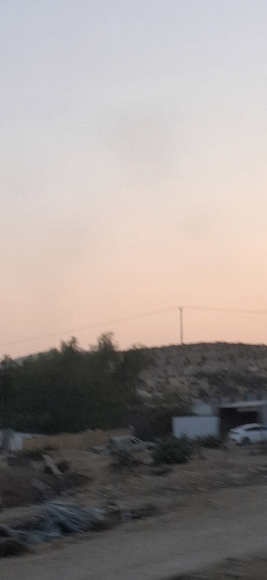 The image captures a twilight scene in Yeruham, Israel, focusing on a semi-arid landscape. The sky displays a soft gradient of pastel colors, transitioning from pale blue at the top to a peachy hue near the horizon. In the middle ground, a dense line of dark green trees forms a stark contrast against a lighter, rough-textured hill that appears to be a man-made mound or a natural geological formation. A tall, thin pole, possibly a utility pole, stands silhouetted against the sky in the upper right portion of the frame.
Below the trees and hill, the foreground reveals a construction or industrial area. Several low-lying structures, including a white rectangular building, are visible. A white car is parked near one of the buildings on the right side of the image. The ground is uneven, covered with dirt, rocks, and some debris, including what appears to be bundles of metal or pipes in the lower left quadrant. The overall impression is one of a developing or industrial fringe on the outskirts of the city, with the natural beauty of the twilight sky providing a serene backdrop.