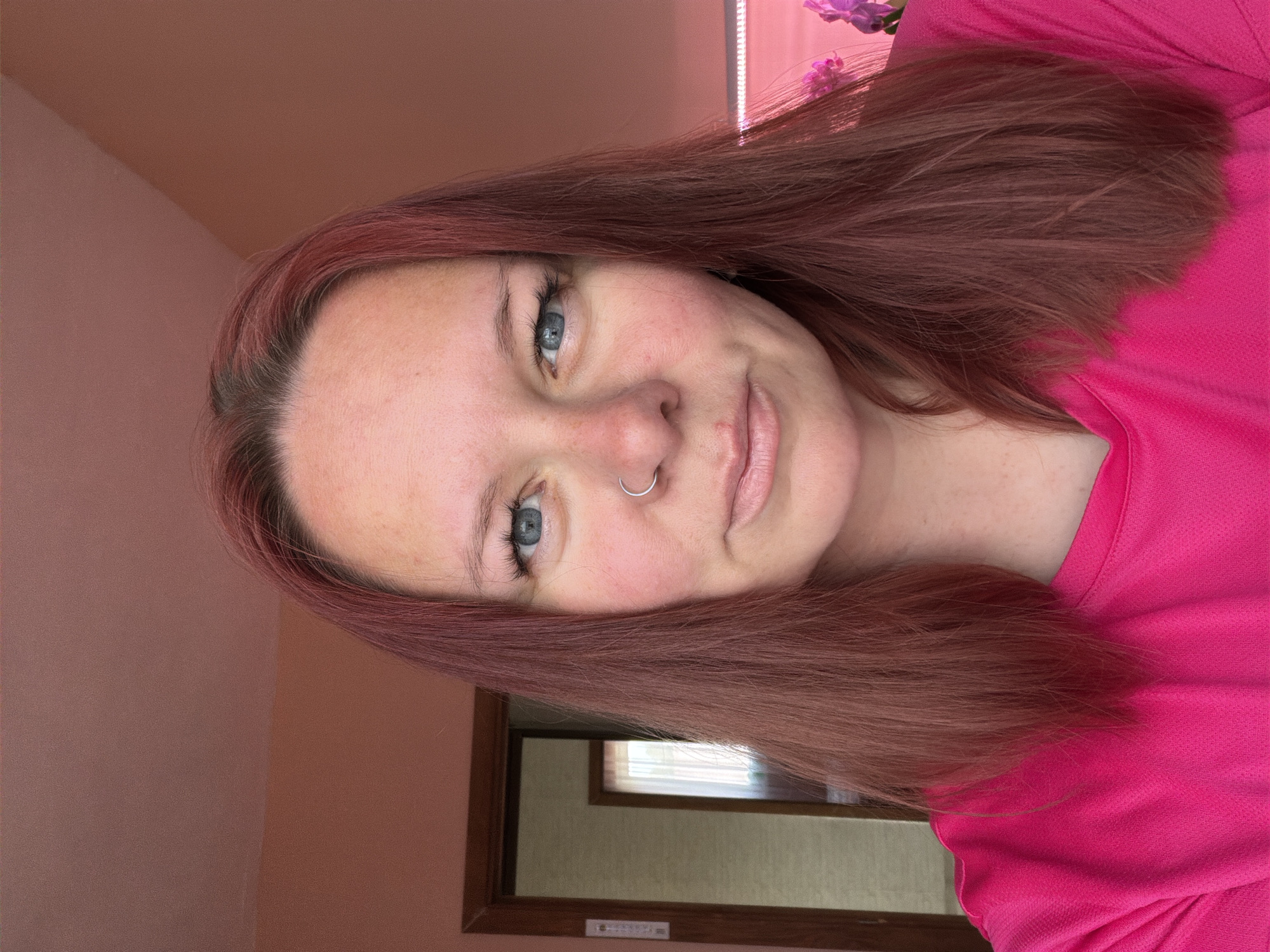 A frontal, close-up portrait of a woman with fair skin and red-auburn hair. She has light blue eyes and a silver nose ring in her left nostril. She wears a bright pink shirt. The background is a peach-colored wall. There is a window and a floral arrangement visible, also illuminated by a fluorescent light. There is no indication that the photo was taken in the city of Chaikovsky, Russia.