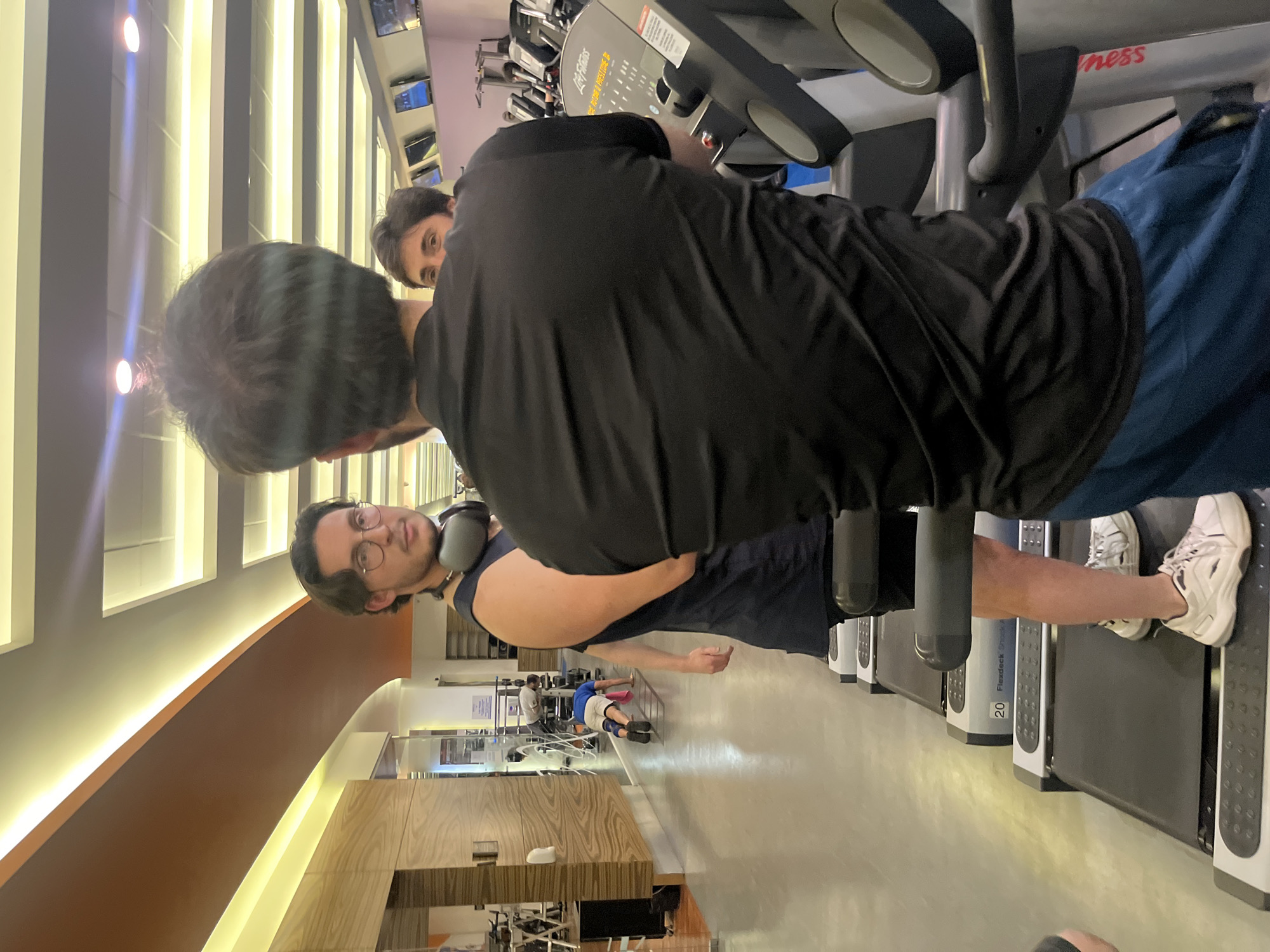 The image depicts an indoor gym facility in Mexico City, Mexico. In the foreground, an adult male is seen from behind, wearing a black short-sleeved shirt and blue shorts, standing on a "Life Fitness" treadmill. Facing him, on an adjacent "Flexdeck" treadmill, is another adult male with dark, wavy hair, eyeglasses, and a dark tank top, with gray headphones resting on his neck; he is looking towards the individual in the foreground. A third adult male's face is partially visible behind the first male's shoulder. In the distant background, a fourth individual wearing a blue t-shirt, light-colored shorts, and knee pads is performing a plank or similar floor exercise, with a pink towel positioned nearby. The gym environment features rows of treadmills, other fitness equipment, wood-patterned wall paneling, a light-colored floor, and several wall-mounted televisions displaying various screens. The ceiling incorporates linear and rectangular lighting fixtures, illuminating the modern fitness center.