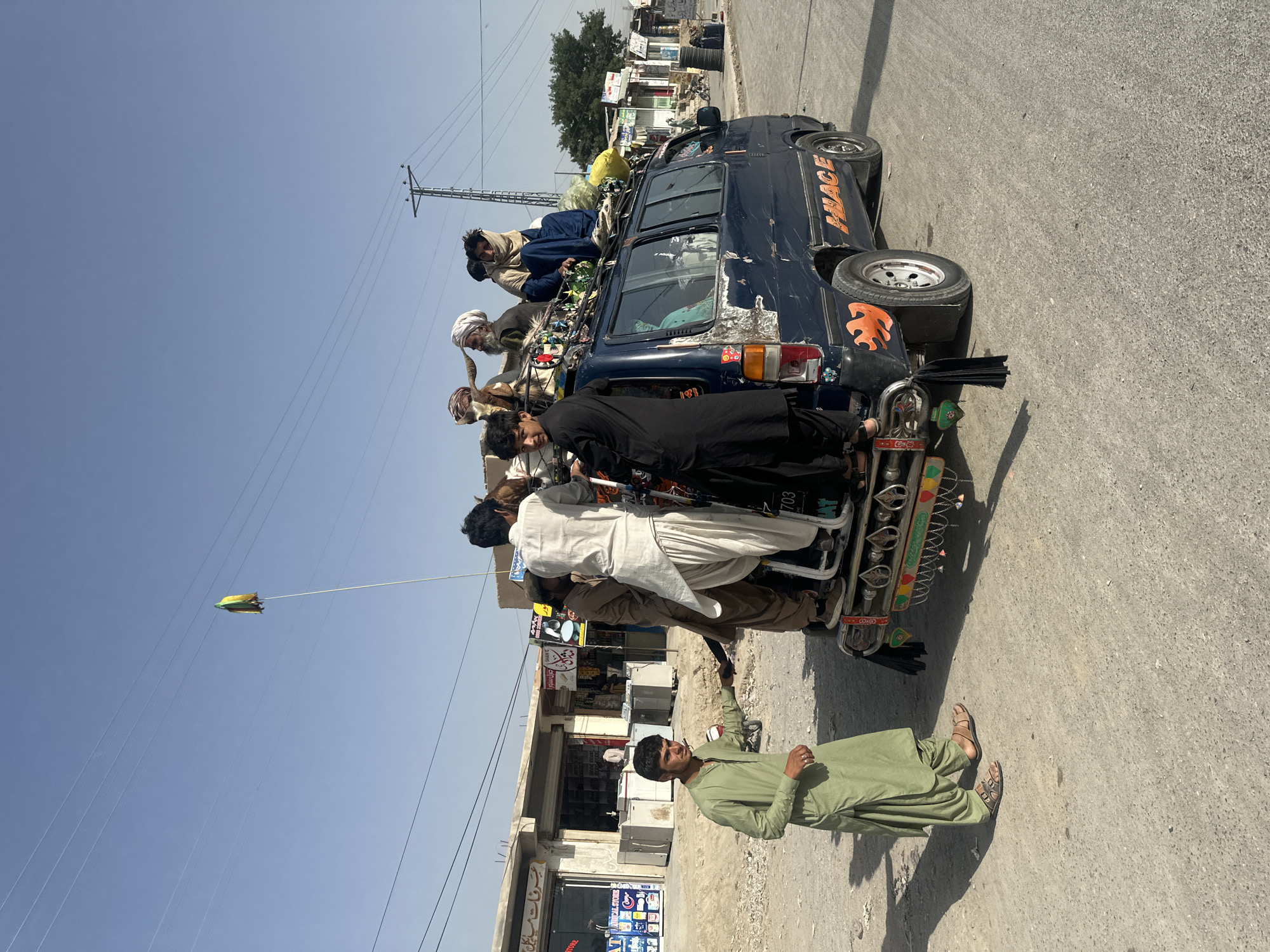 The image depicts an overloaded blue van in Quetta City Tehsil, Pakistan. Several men are riding on the roof and clinging to the rear of the van. The van appears to be modified and decorated. A young man in a green shalwar kameez stands on the street nearby. Buildings and power lines are visible in the background. The scene is typical of transportation practices in some areas.