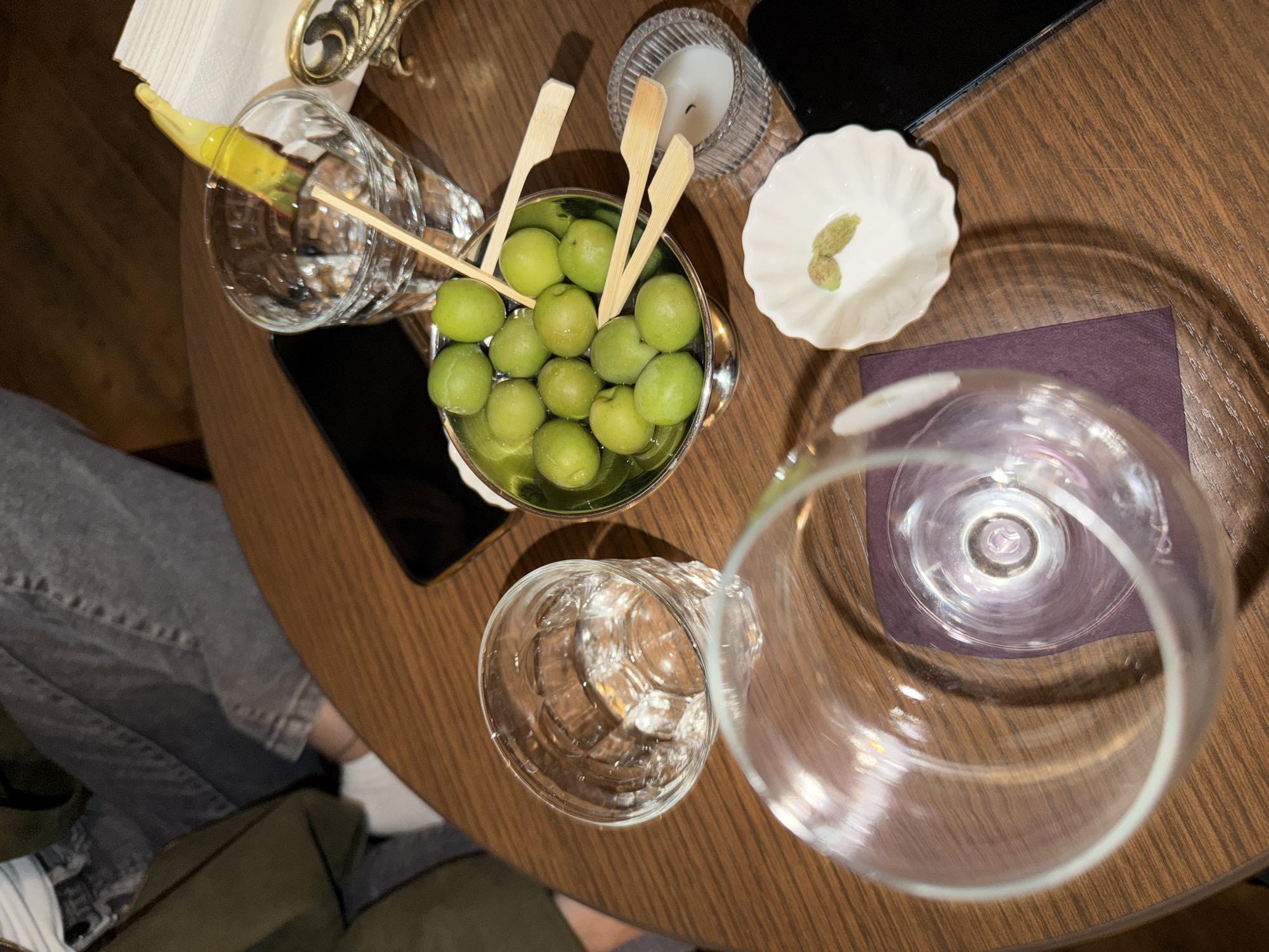 A high-angle shot captures a table in Yerevan, Armenia, featuring culinary and bar items. Central to the composition is a silver bowl filled with green olives, skewered with multiple wooden toothpicks. Surrounding the olive bowl are several clear glass containers: one filled with a clear liquid and adorned with a yellow fruit peel, and two empty glasses. A small white dish contains a couple of small, green seed-like items, adjacent to a lit white candle in a glass holder. A dark-colored device (likely a phone) sits near the candle, and a small purple napkin is also present. Part of a person's clothing is visible in the bottom-left corner. The table itself is made of polished wood, adding to the scene's context.