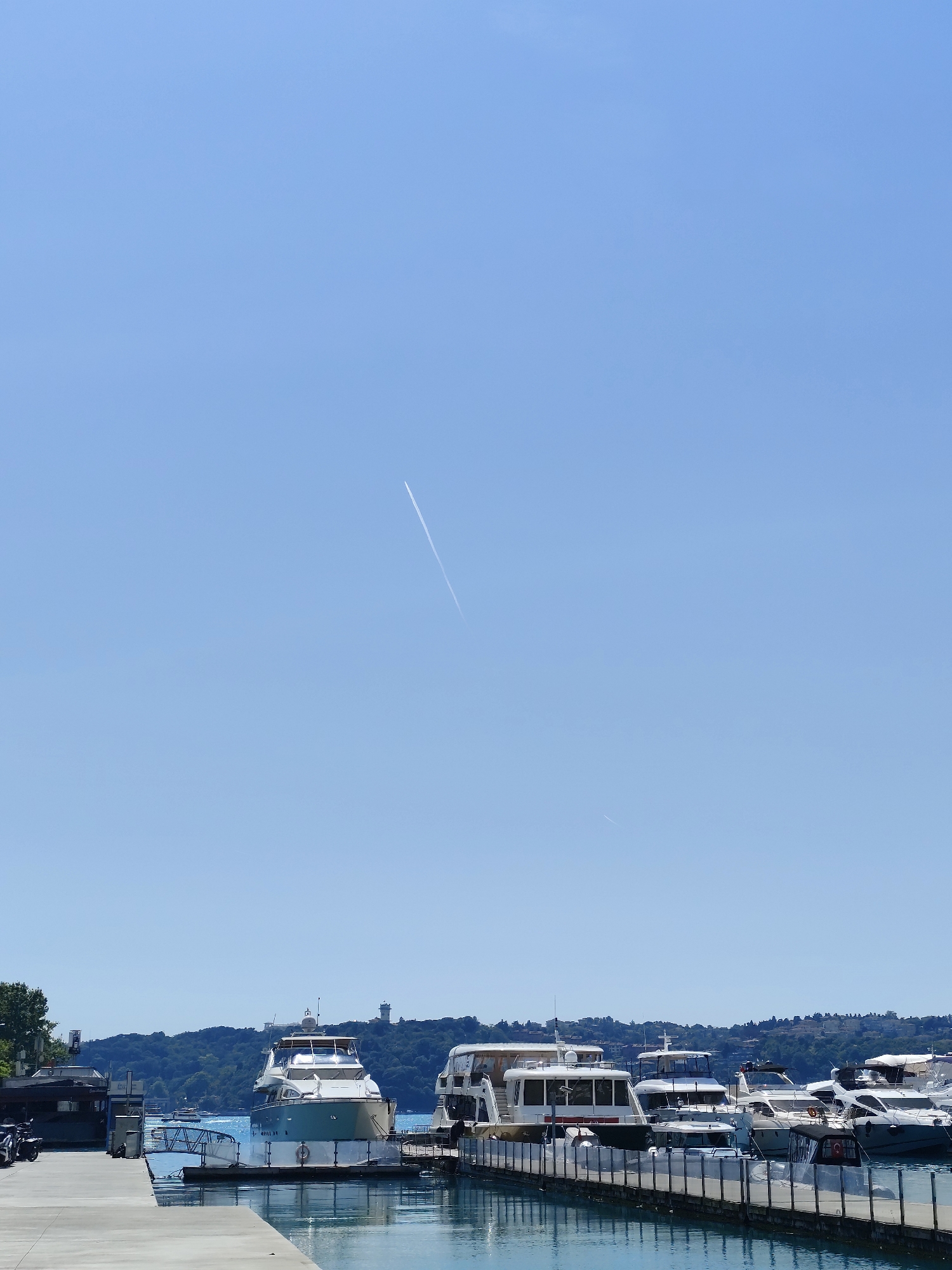 The image captures a vibrant, sunny day at a marina in Sarıyer, Turkey. The upper two-thirds of the frame are dominated by a clear, bright blue sky, traversed by two airplane contrails; one is prominent and extends diagonally from the upper right, while a fainter, shorter contrail is visible further below it.
In the foreground, a concrete pier extends from the left, where some dark objects, possibly motorcycles or scooters, are parked near a small, dark-roofed building. To the right, another concrete pier with white metal railings supports numerous boats. The marina itself is bustling with moored vessels. A large, well-maintained yacht with a light-colored hull and distinctive green-blue accents is prominently docked on the left side of the marina area. To its right, a variety of other motor yachts and smaller boats are closely clustered, featuring different designs, some with enclosed cabins and others with open decks. The water in the marina is calm and reflects the clear sky and boats.
The background consists of a lush, verdant hillside covered densely with trees and some scattered buildings among the foliage. A distinct white, tower-like structure, possibly a lighthouse or communication tower, stands out on the distant hillside. The scene is brightly lit by natural sunlight, indicating a mid-day setting with excellent, clear weather. No people are visible in the frame, and there is no discernible text on any of the boats or structures. The overall impression is one of a peaceful and picturesque coastal or lakeside environment.