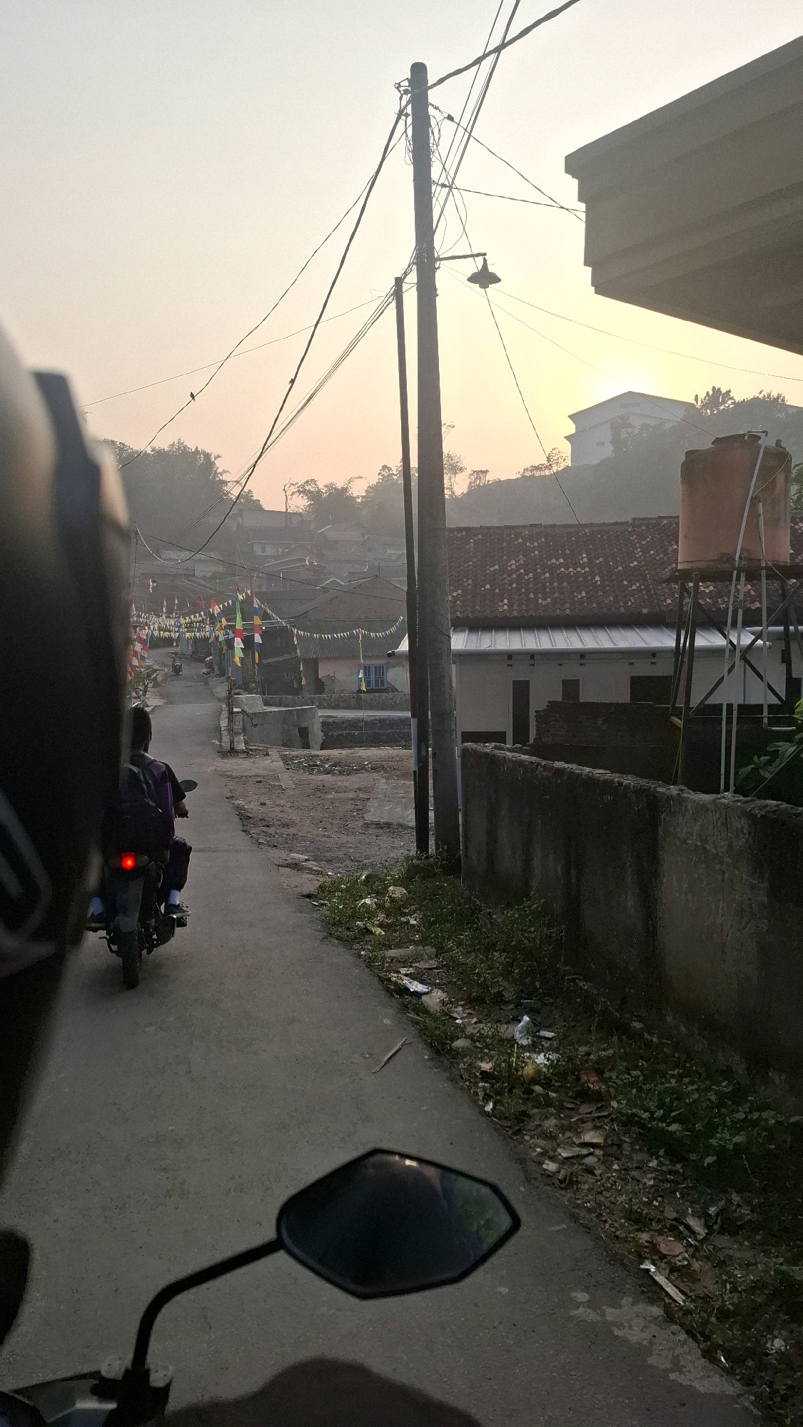 The image depicts a scene from a moving vehicle, likely a motorcycle, on a paved road in a rural or suburban area of Bandar Lampung, Indonesia. The time of day appears to be early morning or late afternoon, with a soft, hazy light illuminating the sky and the landscape.
In the foreground, the handlebars and a mirror of the motorcycle are visible, suggesting the perspective of the rider or a passenger. A person wearing a purple shirt and dark pants, with a backpack, is riding on a motorcycle ahead of the viewpoint vehicle. This person is heading away from the viewer, down the road.
The road itself is a paved asphalt surface, with a slight incline leading into the distance. On either side of the road are houses and buildings with tiled roofs. Many of these buildings are adorned with colorful flags and pennants, suggesting a festive occasion or a local celebration. Trees and vegetation are visible in the background, adding to the natural landscape. A tall water tank stands on a metal structure near one of the buildings.
Utility poles with numerous wires crisscross the scene, indicating the presence of electricity and telecommunication services. The sky is overcast with a warm, diffused light, creating a tranquil and somewhat muted atmosphere. There is some litter visible along the side of the road. The overall impression is one of a quiet, everyday scene in a community setting.