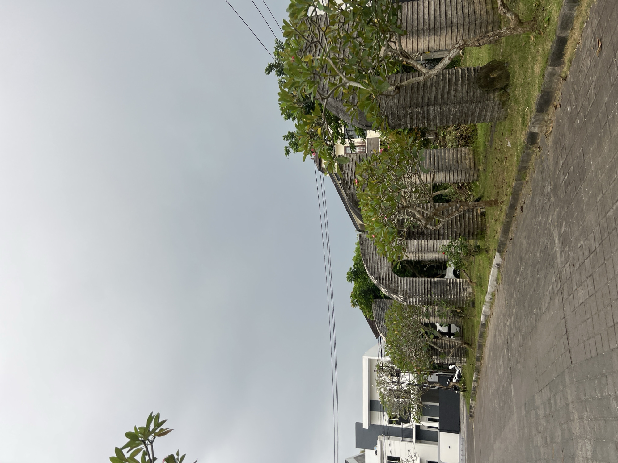 The scene shows a streetscape in Kuta Selatan, Indonesia, with a unique architectural feature along the roadside. A structure composed of stacked, horizontal, concrete-like slabs creates an arched or undulating wall, seemingly part of a property boundary. The wall is partially overgrown with grass and small trees, giving it a weathered or aged appearance. Adjacent to this structure is a paved street or walkway made of interlocking gray blocks. Further along the street, a more modern, two-story building is visible, with a white facade and contrasting gray or black architectural elements. The sky is overcast, with a uniform gray color, suggesting a cloudy day. Power lines are strung overhead. The photograph appears to be taken at ground level, looking slightly upward toward the structures and sky. The orientation of the image is rotated counter-clockwise.