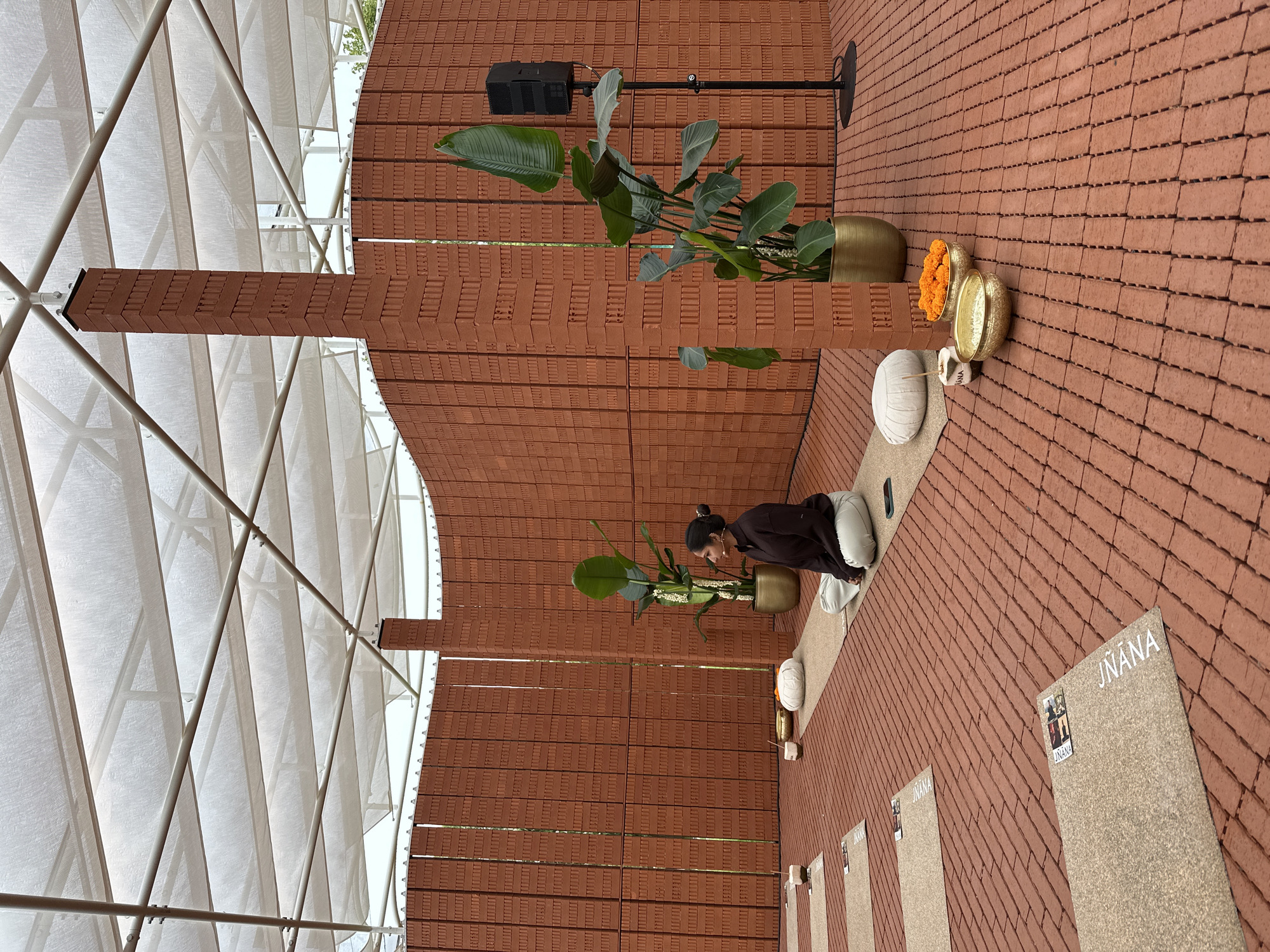 This image depicts an outdoor meditation space, likely in London, United Kingdom, constructed within a pavilion-like structure with a translucent white roof supported by a metal framework. The walls and floor are made of orange-red bricks. A woman is seated in a cross-legged meditation pose on a mat. In front of her, there are meditation cushions and golden bowls containing orange marigold flowers. To her right, a large potted plant with broad green leaves stands next to a speaker on a stand. Several other meditation mats are visible on the floor, some bearing the word "JNANA". The environment appears serene and designed for mindfulness practices.
