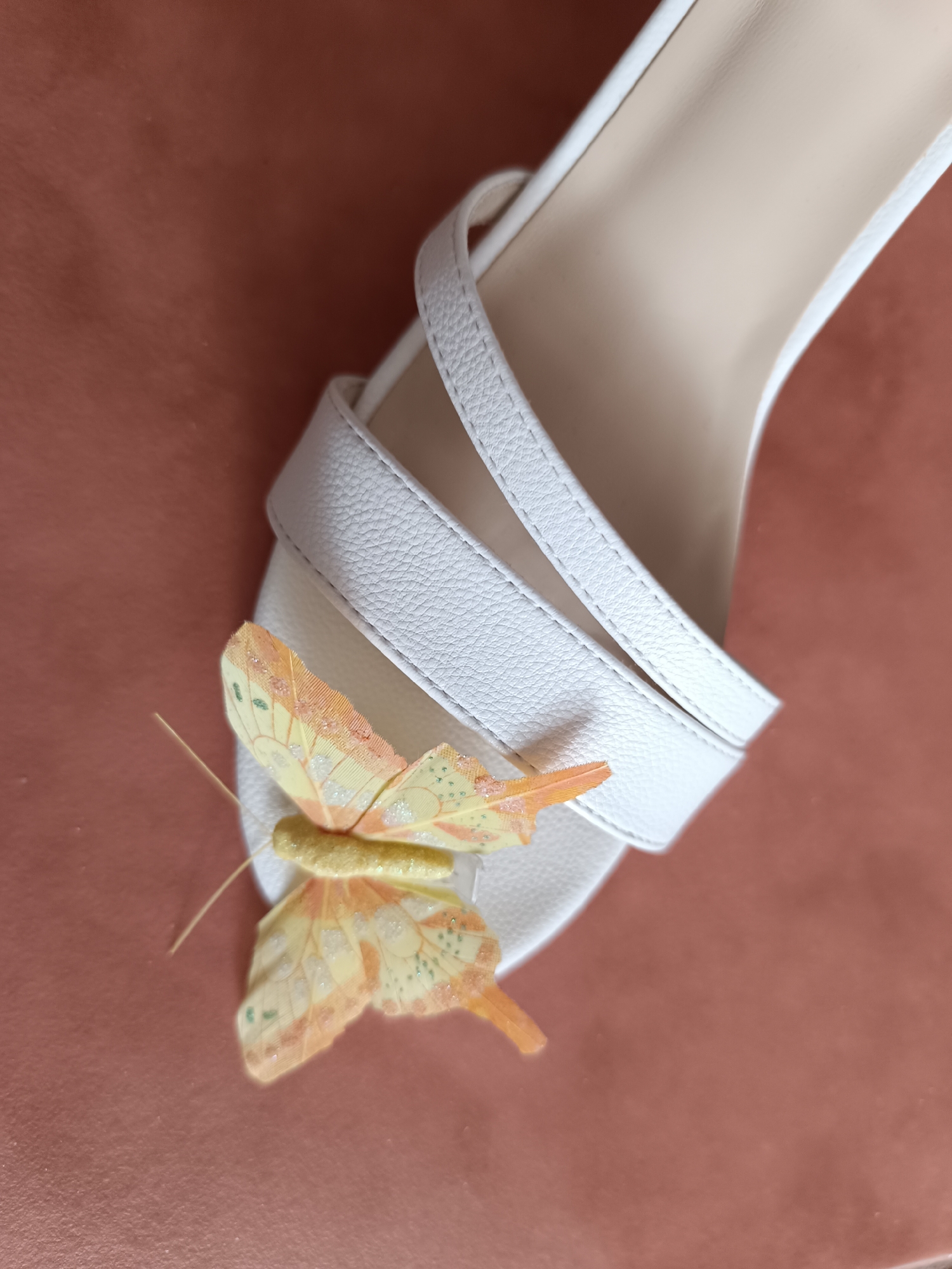 The image shows a close-up of a white sandal with a decorative yellow butterfly. The sandal is made of textured leather and has thin straps. The butterfly is positioned on the front strap of the sandal. The background is a muted reddish-brown surface. The image is taken from a high angle, looking down at the sandal. There is no information available to indicate the location in Ternopil, Ukraine.