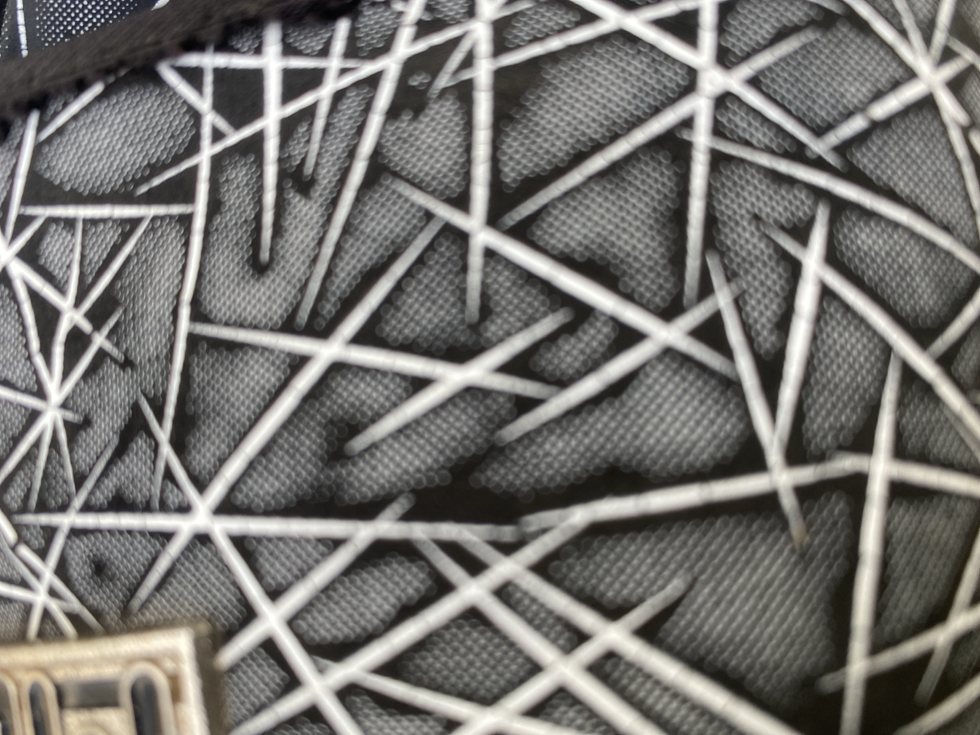 The image shows a close-up, blurry view of a black fabric with a white geometric pattern. The pattern consists of intersecting lines that create a web-like or fragmented star-like design. The fabric beneath the pattern appears to have a textured, dotted surface. A small portion of a light-colored, rectangular object with dark outlines is visible on the far left of the image. To the right, a dark, ribbed edge of another object is partially visible. The scene is indoors or in a protected outdoor space, with no visible environmental context for Jalingo, Nigeria. There are no people or discernible actions taking place.