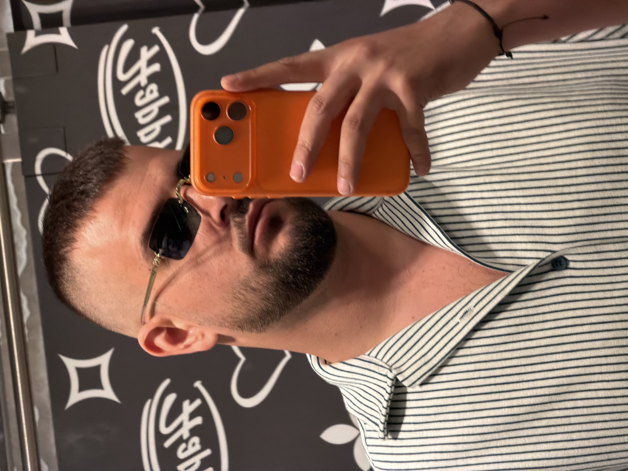 A man with short dark hair and a beard takes a selfie using an orange-colored phone. He is wearing sunglasses with gold frames and a striped shirt. The background appears to be a dark surface with a white logo. The specific location in Plovdiv, Bulgaria is not discernible from the provided image.