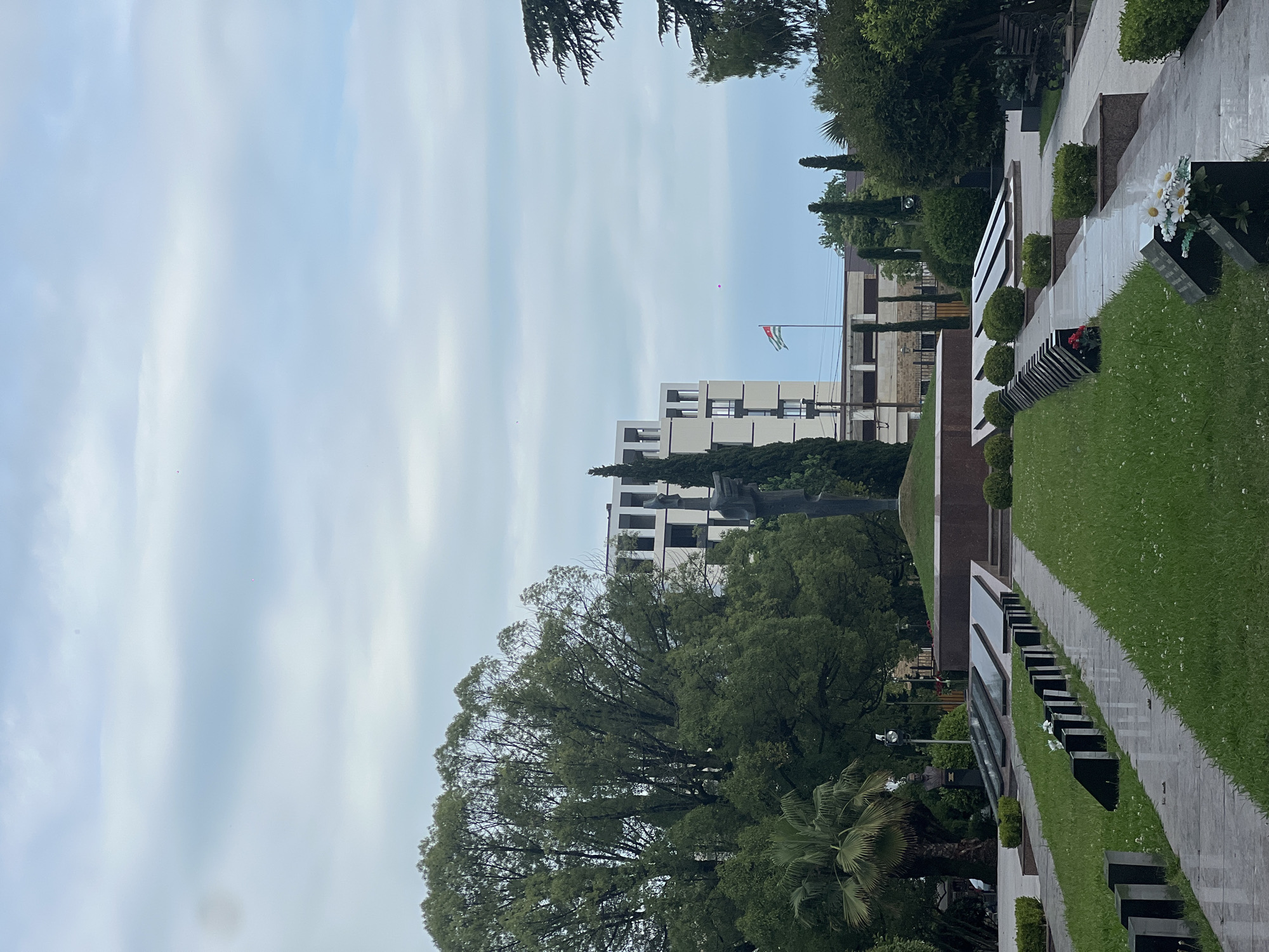 The image shows a memorial park with a flag flying in the background. The flag is green, white, and red with an emblem in the center. Behind the flag, there is a multi-story building with a modern architectural style. In the foreground, there are rows of black granite headstones or memorial markers on a manicured lawn. A paved walkway runs alongside the headstones. There are also several well-maintained bushes and trees, including a large leafy tree on the left side of the frame and palm trees on the right. Artificial white daisies are placed on one of the memorial markers. The sky is overcast with light blue and white clouds, suggesting it might be a cloudy day. The overall scene appears to be solemn and peaceful, typical of a memorial or cemetery. There is no visible text in the image.