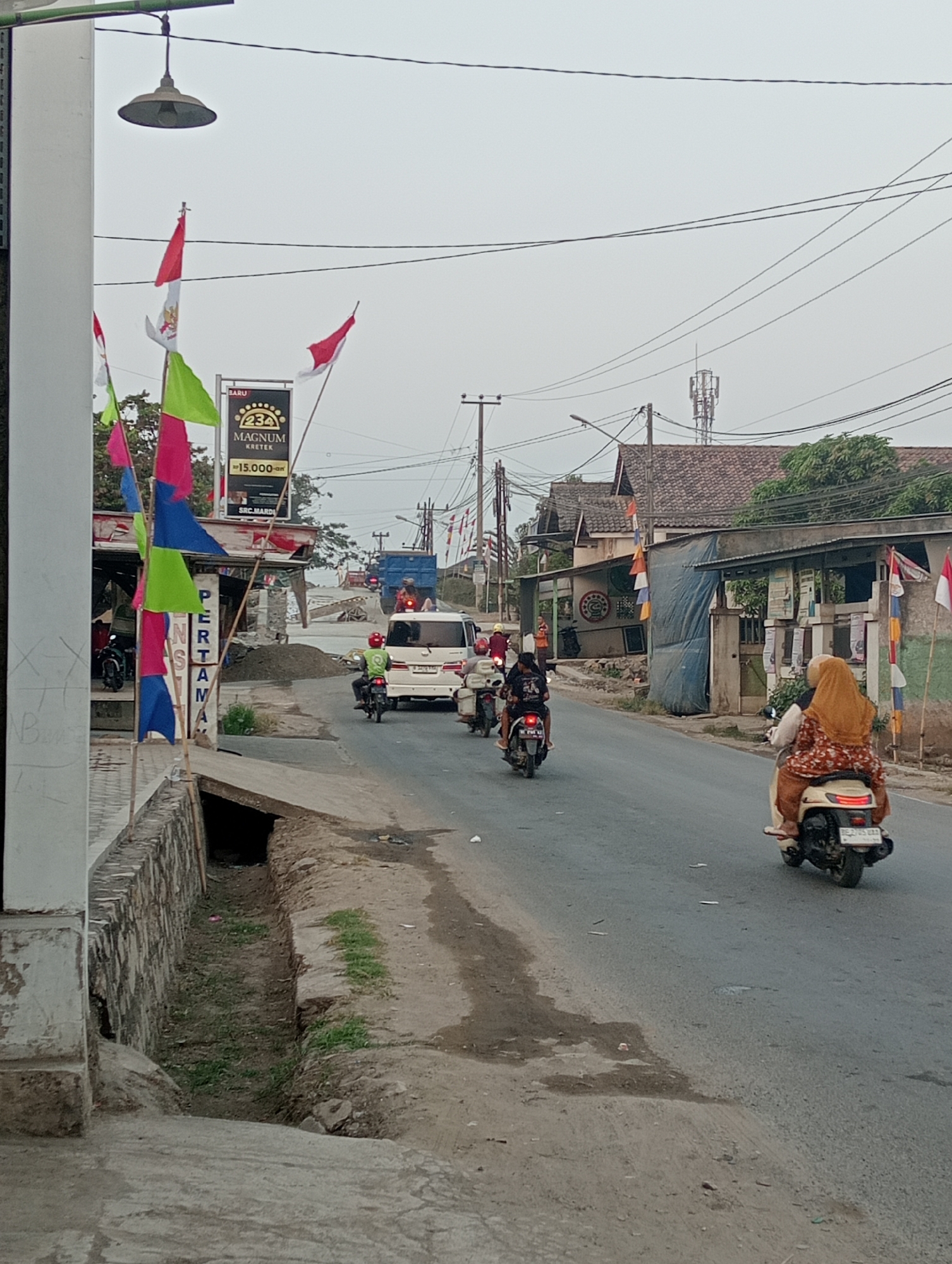 The image depicts a street scene in Pringsewu, Indonesia. The sky is overcast, suggesting a cloudy day. The road is paved, with a concrete drain on the left side of the image. Along the side of the road, there are various structures, including what appears to be a small shop with a sign advertising "MAGNUM KRETEK" and a price of "Rp15.000,-". There are also several Indonesian flags, predominantly red and white, and some colorful pennants displayed.
Traffic is present on the road, consisting of several motorbikes and a white van. On the motorbikes, there are multiple individuals, some wearing helmets, including a woman in the foreground riding a scooter with a passenger. The woman is wearing a headscarf and a patterned dress. The people on the motorbikes are in motion, suggesting they are traveling along the road.
In the background, there are buildings with tiled roofs and vegetation. A cell tower is visible in the distance. The overall impression is a typical roadside scene in a semi-urban or rural area of Indonesia, with elements of commerce, transportation, and local decorations.