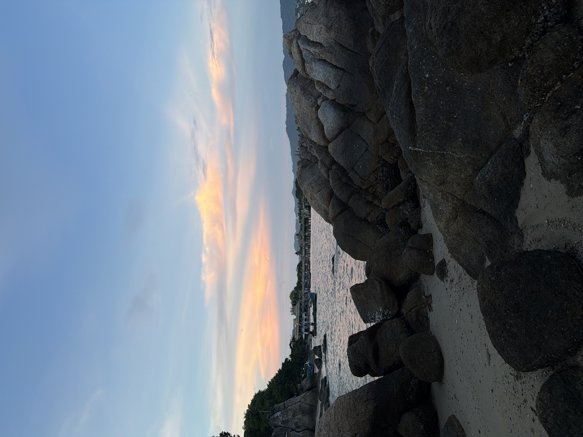 The image depicts a serene coastal scene during sunset in Ko Samui District, Thailand. The sky is a gradient of soft blues and pastel pinks, with wisps of clouds catching the last rays of the sun. In the foreground, large, dark, weathered boulders lie scattered on a sandy beach, some partially submerged in the gentle, rippling water. Beyond the immediate shoreline, the sea stretches towards a distant, hazy coastline dotted with buildings and a pier. Several small boats, including a longtail boat with people aboard, are visible on the water, suggesting a tranquil fishing or leisure activity. Lush green trees and foliage frame the left side of the image, adding a touch of natural beauty to the coastal landscape. The overall impression is one of peaceful, tropical twilight.