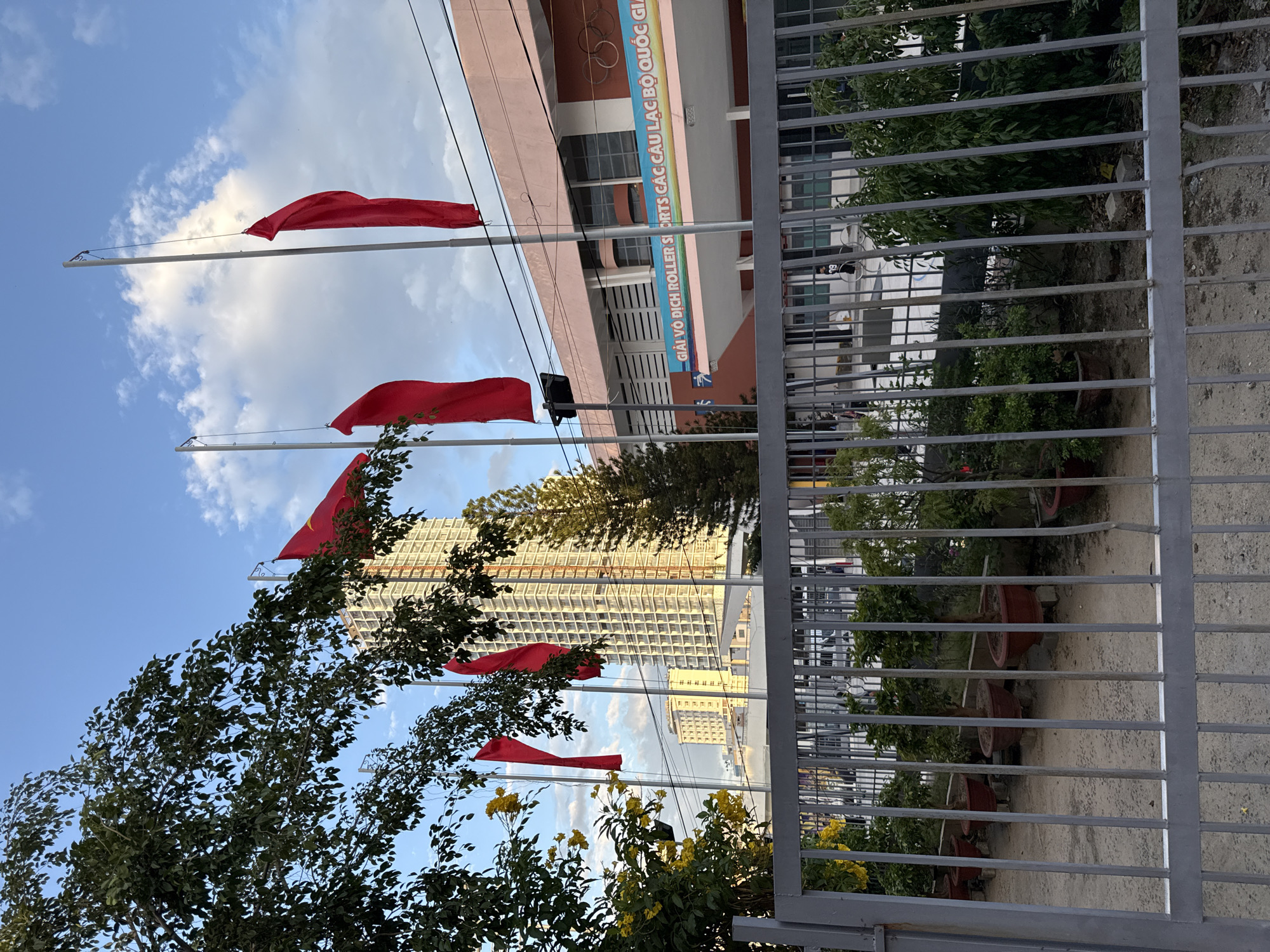 The image depicts a street scene in Nha Trang, Vietnam, dominated by a tall, modern building with a light-colored facade and numerous windows. In the foreground, a gray metal fence encloses a pathway with several potted plants and trees. Red flags flutter from tall poles in front of a pink building with a banner displaying Vietnamese text and what appear to be Olympic rings. The sky is blue with scattered clouds. The overall impression is one of an urban environment with some landscaping and official signage.