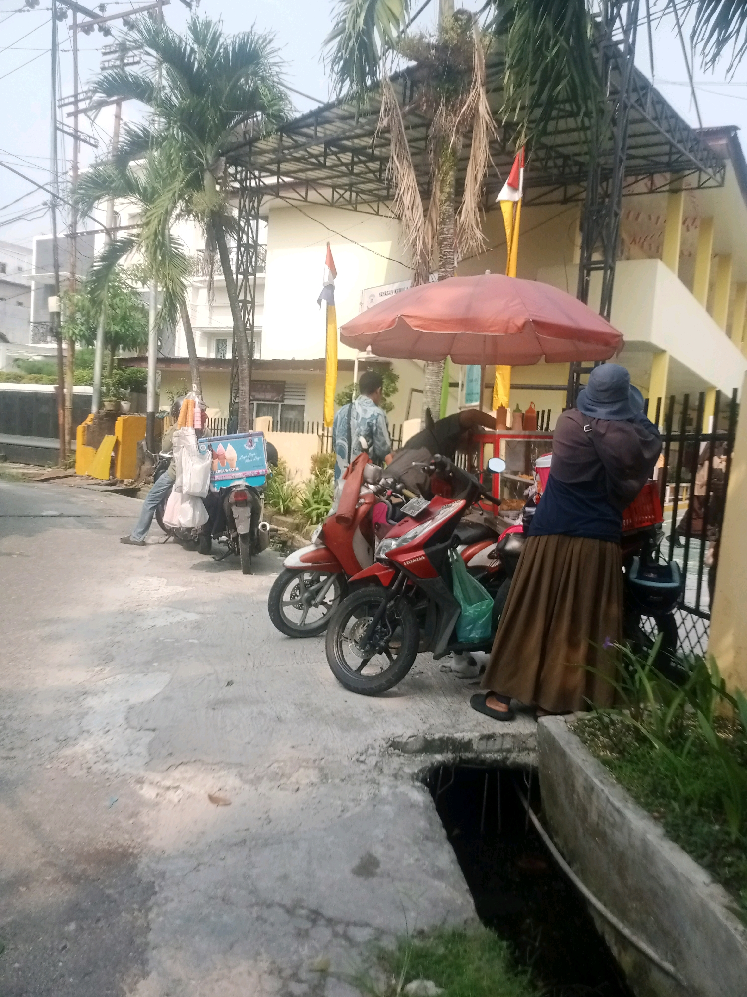 This is a medium-shot, eye-level, outdoor photograph taken during the daytime in Pekanbaru, Indonesia. The scene depicts a street with parked motorcycles and a person selling ice cream from a mobile cart.
In the foreground, the pavement is cracked and worn, with a drainage ditch running along the right side of the image, filled with dark water and some visible debris. Green grass and plants are growing along the edges of the ditch.
Dominating the middle ground are two motorcycles. The one on the left is a dark-colored scooter with an ice cream cooler box mounted on the back, displaying images of ice cream cones and the text "ICE CREAM CONE". Attached to the cooler box are several white plastic bags and what appear to be ice cream cones. The cooler box is blue and white. A person, partially obscured, is seated on this motorcycle, wearing a blue shirt and jeans. The motorcycle on the right is a red and white scooter.
In the background, a woman wearing a blue headscarf and a long brown skirt is standing next to the ice cream cooler box, seemingly interacting with the vendor or observing the scene. She is wearing a brown jacket over a blue shirt. The woman is standing on the right side of the frame. Behind the motorcycles, there's a red umbrella serving as shade for the ice cream vendor. Beyond that, a building with a yellowish exterior and a metal roof structure is visible. Palm trees are present in the scene, adding to the tropical environment. A flagpole with an Indonesian flag is also visible. The sky is overcast.