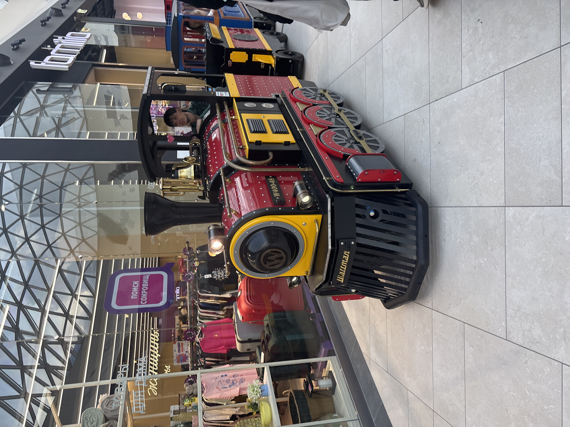 The image depicts an indoor scene within a shopping mall in Perm, Russia, featuring a miniature, multi-car train operating on a light-colored tiled floor. The locomotive, primarily red and black with yellow accents, displays "Wattman" on its front grill and "W400-48" on its side, equipped with a brass bell and a headlight. A young adult male with dark hair, wearing a dark green plaid shirt, is seated as the driver in the train's first car, looking slightly to his left. Behind the driver's car are at least two additional passenger cars; the first is red and black, and the second is blue and black, with a partial figure visible inside. On the far right, the lower body of an individual wearing dark pants and white shoes, holding a white bag, is partially visible, standing adjacent to the train.
The mall environment includes large glass storefronts on both sides. The left storefront displays clothing items, including pink t-shirts and fuchsia blouses on racks, alongside luggage (red, grey, green) in the window. Visible Russian text on signs within this store includes "ПОИСК СОКРОВИЩ" (TREASURE HUNT) and phrases related to "FOR HOME WOMEN". To the right, a store entrance is marked by a prominent white "Familia" sign. The ceiling features a modern, multi-paneled glass and metal grid structure. The floor is composed of large, light grey rectangular tiles with dark grey grout lines.