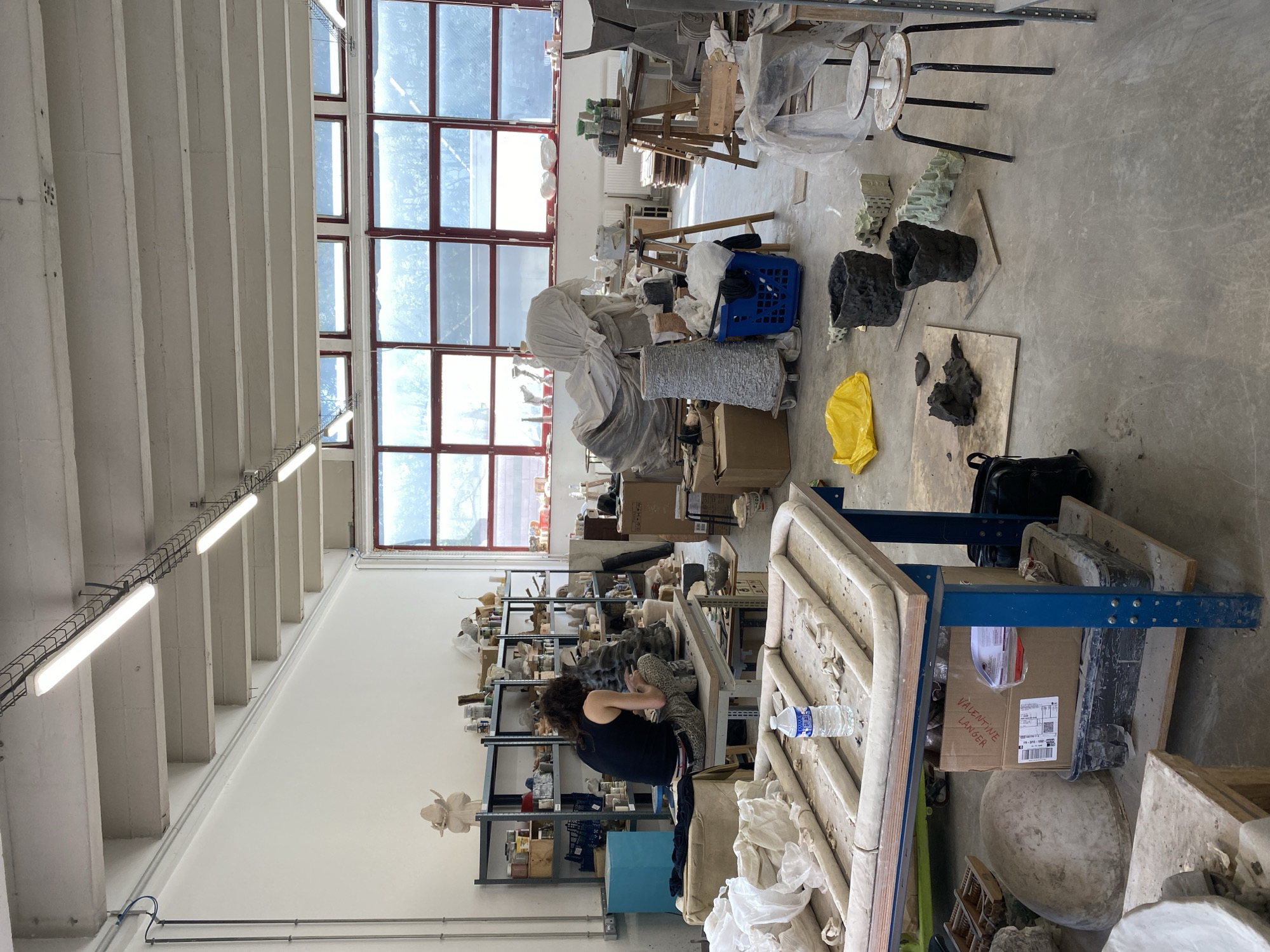 The image, oriented on its side, depicts a bustling art studio or workshop, likely located in Saint-Ouen-sur-Seine, France, filled with an array of sculptures, molds, and artistic materials. The large space is brightly illuminated by natural light pouring through a series of tall, gridded windows with reddish-brown frames, set into a white wall. The ceiling features exposed concrete beams and long rows of fluorescent lighting fixtures.
In the mid-ground, a woman with dark, curly hair, wearing a black tank top and patterned trousers, is seated at a workbench, intently engaged in working on a grey, textured sculpture. Behind her, metal shelving units are densely packed with numerous smaller art pieces, molds, and studio supplies.
The concrete floor is cluttered with various art projects and tools. Visible items include several dark, organic-shaped sculptures, lighter, intricately textured forms, a blue plastic basket, and a bright yellow plastic bag. Further into the background, multiple worktables are laden with additional art in progress, some covered with white sheets.
Notable text includes the handwritten name "VALENTINE LANGER" clearly visible on the side of a large cardboard box in the lower right portion of the image. A plastic water bottle is also present near this box. The scene conveys an active and industrious artistic environment, characterized by ongoing creative production.
