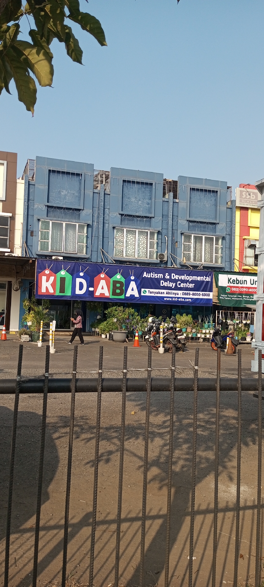 In the city of Lambangjaya, Indonesia, a blue, multi-story building stands prominently. A large banner across the lower part of the building advertises "KID-ABA Autism & Developmental Delay Center." To the right of this banner, another sign indicates "Kebun Ur." and displays a phone number and text mentioning plants. In front of the building, a person is walking, and several motorcycles are parked. The scene is captured from behind a metal fence, with tree shadows cast upon the ground.