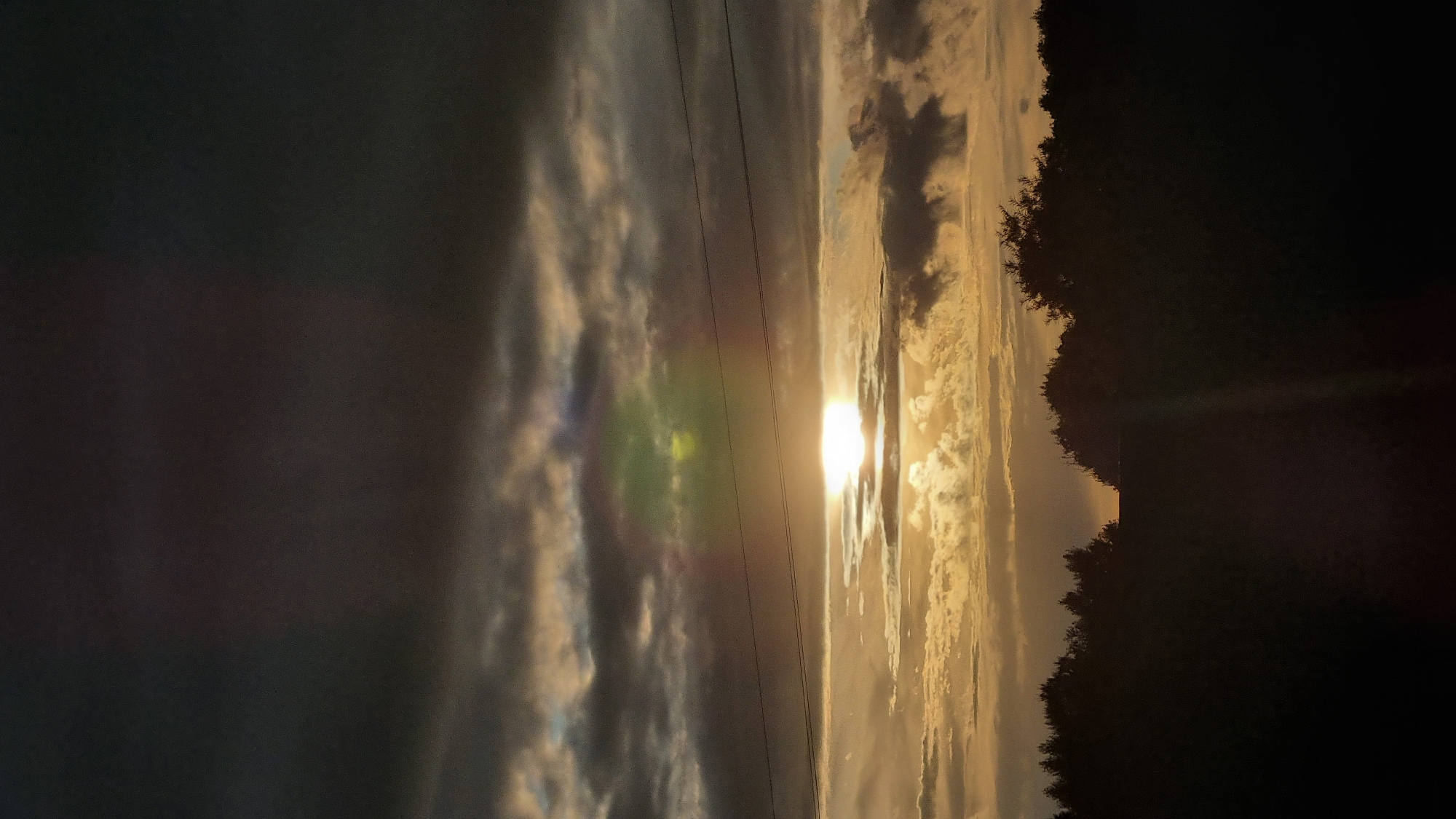 The image depicts a sunset over a silhouetted tree line in Vysokoe, Russia. The sky is filled with dramatic, dark clouds interspersed with streaks of golden light from the setting sun. Two thin power lines are visible stretching horizontally across the middle of the frame. A faint rainbow-like lens flare is present in the upper portion of the sky. The lower portion of the image is dominated by the dark silhouette of trees, obscuring any details of the landscape or any people or objects within Vysokoe. The overall impression is one of a moody and atmospheric natural scene.
