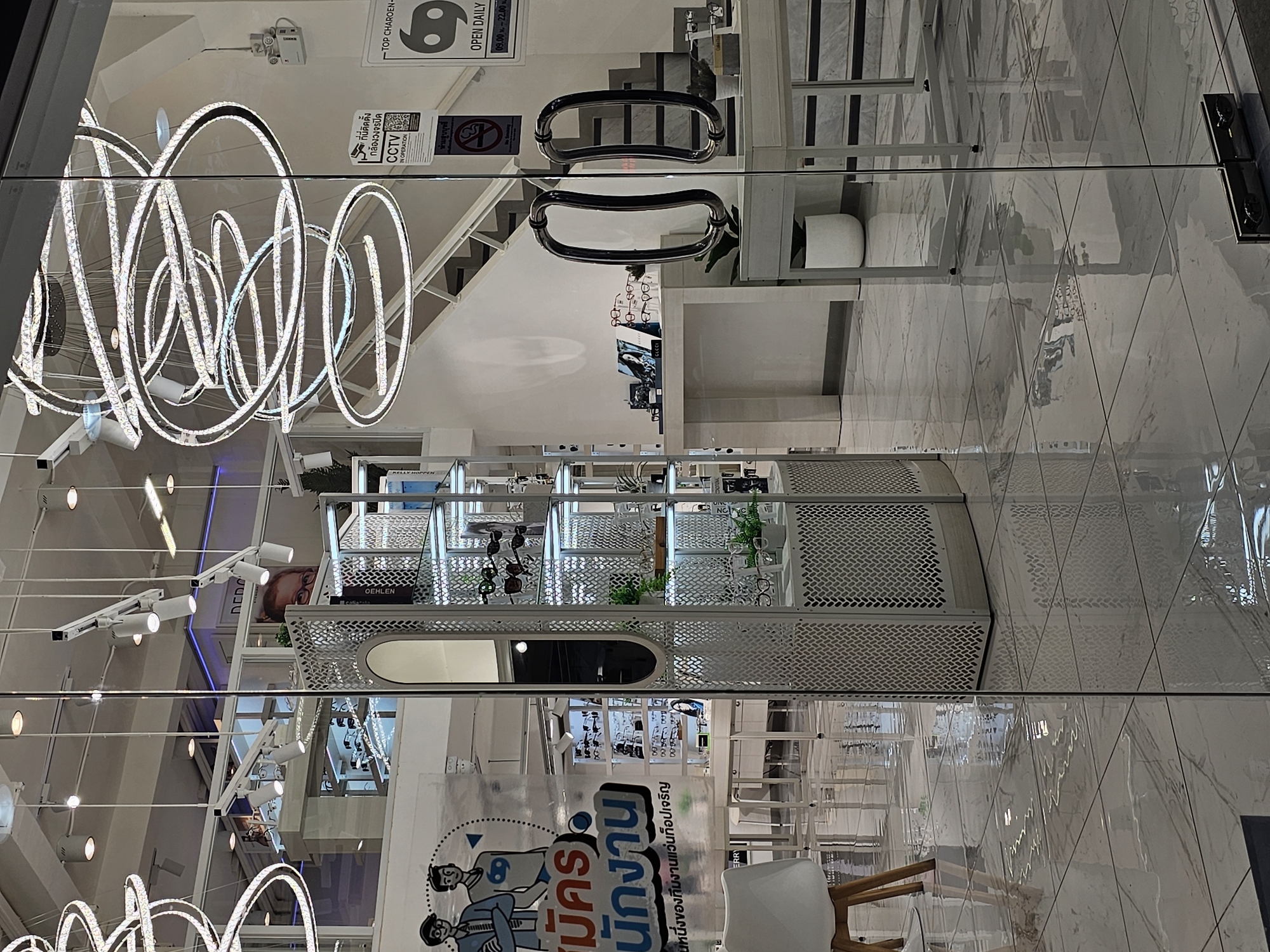 The image depicts the entrance of an optical shop in Bangkok, Thailand. The shop's interior is visible through a glass door, revealing a clean, modern design with white and metallic accents. Suspended from the ceiling are elaborate, multi-ringed chandeliers that emit a bright, white light. Displays of eyeglasses are showcased on shelves and tables. In the background, a staircase leads to an upper level. Several signs are visible, including a CCTV notice and operational hours. The floor is made of polished marble tiles, which reflect the shop's lighting and interior elements. The exterior area appears wet, possibly from recent rain, creating further reflections on the floor. The overall impression is that of a well-lit, organized retail space.