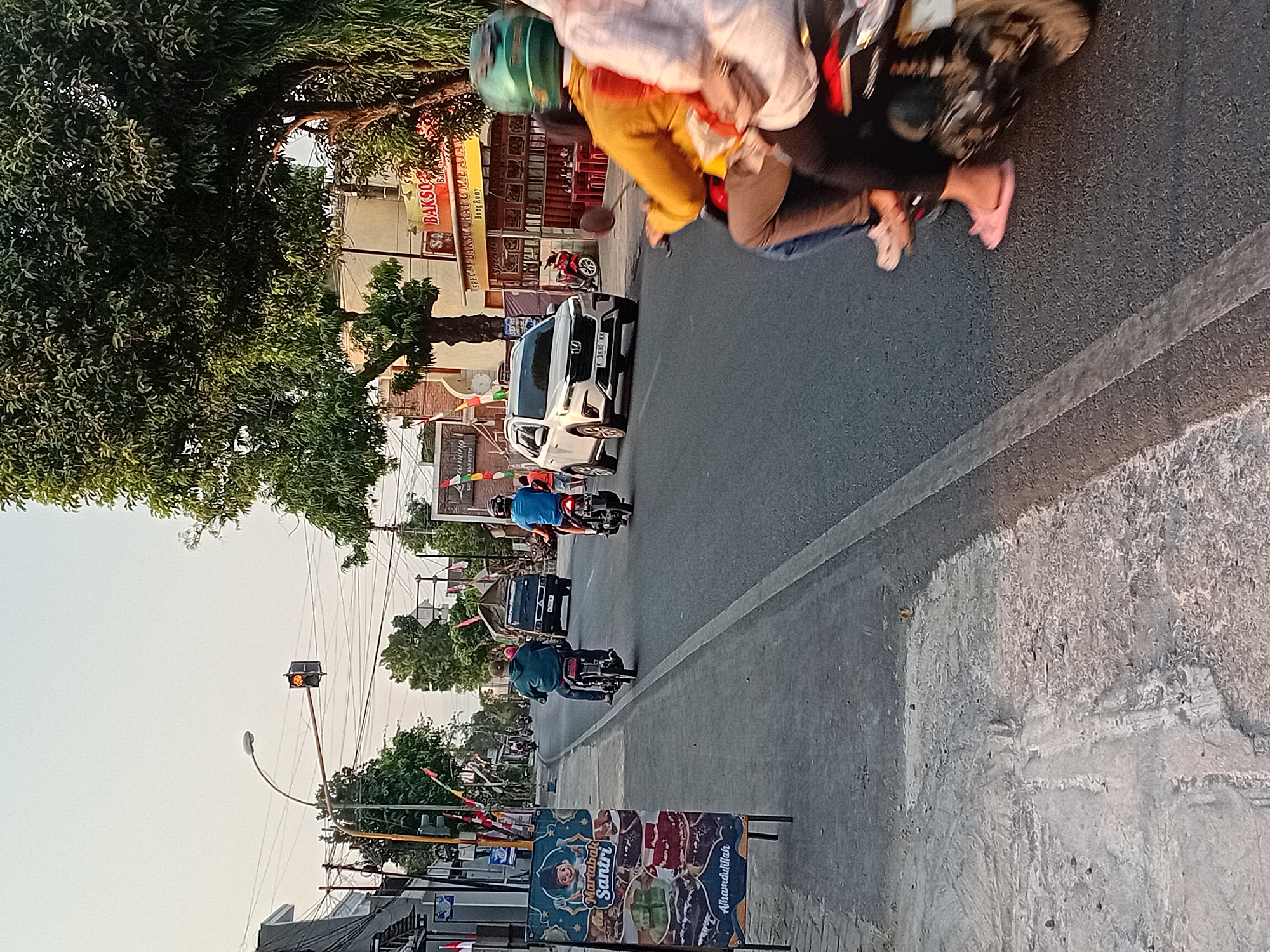 In Damaran, Indonesia, a street scene unfolds with traffic and pedestrian activity. A traffic light at an intersection shows a yellow light, indicating a transition between red and green. On the sidewalk, a sign displays images of various food items, including cakes and sweet treats, with the text "Mariabak Santri" and "Alhamdulillah". Further down the street, a white SUV drives on the right side of the road, while several motorcycles are present. Two individuals are riding on a motorcycle in the foreground, one wearing a yellow top and the other in a light-colored shirt, both with helmets. Another motorcycle with a rider in a blue shirt is further down the road. Buildings line the street, some featuring storefronts with signage, including one that reads "BAKSO". Trees with lush green foliage are visible, casting shadows on the pavement. The sky is clear, suggesting a daytime scene, possibly late afternoon due to the lighting. The road surface shows a white line marking the lane division.