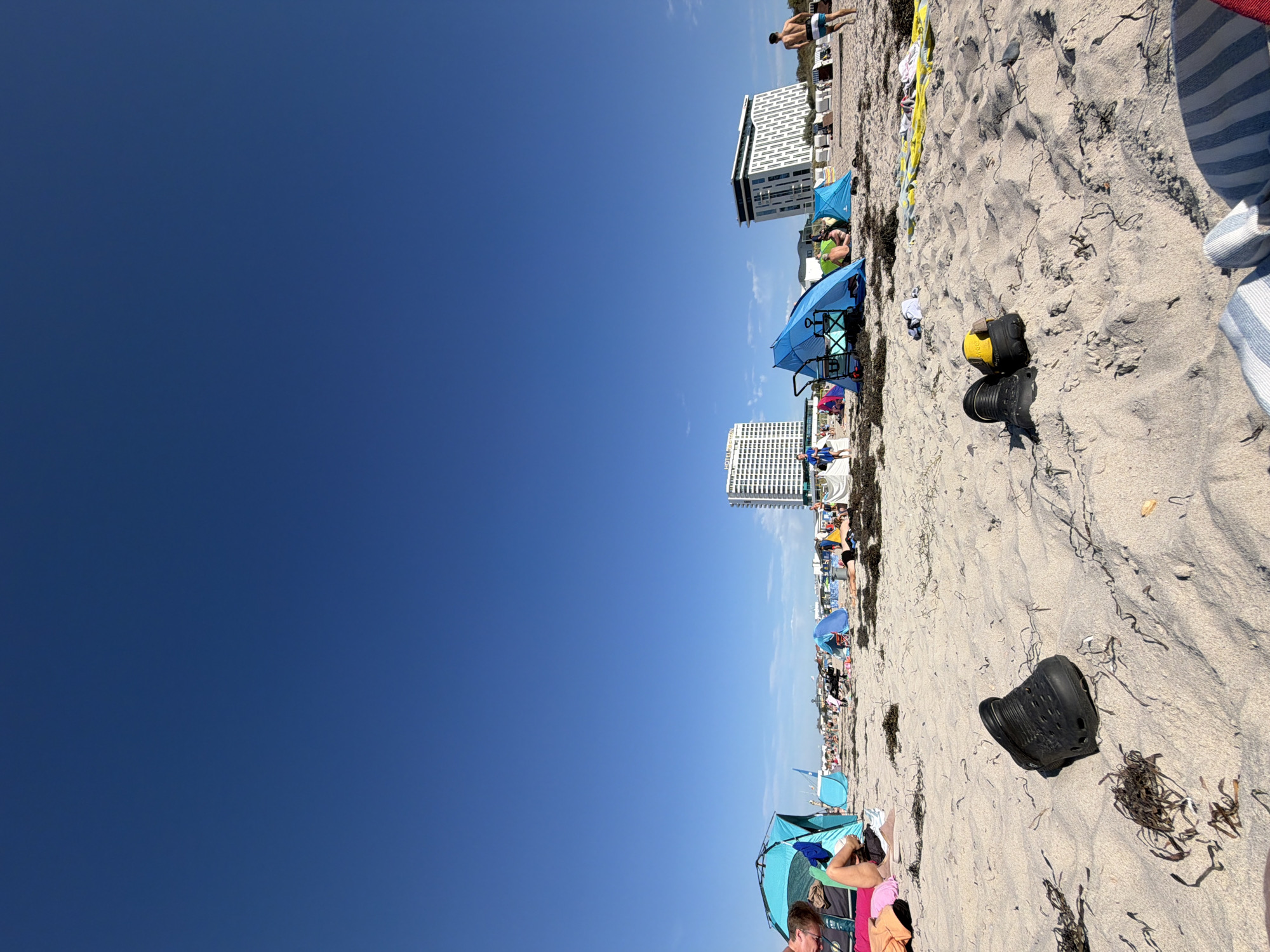 The image depicts a sunny day at the beach in Warnemünde, a seaside resort district of Rostock, Germany. The foreground is dominated by a sandy beach with scattered clumps of seaweed. Two black clogs are prominently placed in the sand, with one of them slightly isolated. In the middle ground, several beach tents, including a bright blue one with a pull-cart, are set up. People are visible relaxing on the beach and walking around. In the background, a large, modern white hotel building stands tall against a clear blue sky. Another contemporary building with a dark facade and a white patterned exterior is also visible to the right of the hotel. A few beach chairs and umbrellas can be seen further down the beach. The scene conveys a sense of leisure and summer recreation.