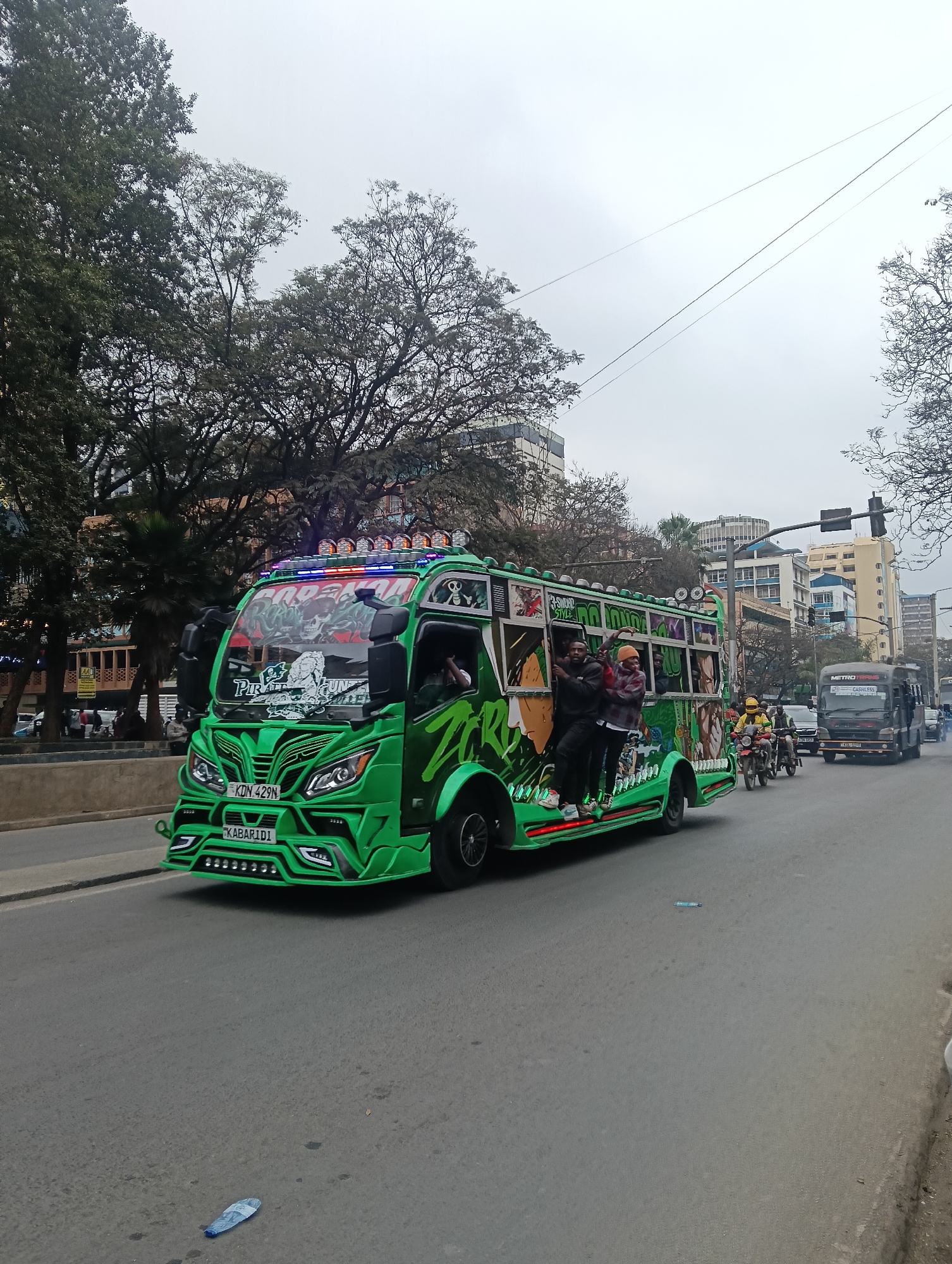 This image captures a vibrant green matatu, a form of public transportation common in Nairobi, Kenya, driving down a city street. The matatu is elaborately decorated with colorful graffiti, lights, and imagery, including skulls and what appears to be a pirate theme on the windshield. Several passengers are visible, some standing and leaning out of the windows, suggesting a lively atmosphere.
The street appears to be a multi-lane road in an urban setting. In the background, buildings of varying heights and architectural styles are visible, indicating a developed city environment. Trees line the side of the road, and power lines are strung overhead. Other vehicles, including a black bus with "METRO TRANS" and "CASHLESS" written on its side, and motorcycles, are also present on the road, suggesting typical city traffic.
The overall impression is that of a bustling day in Nairobi, with the highly customized matatu being a focal point. The sky is overcast, suggesting a cloudy or potentially rainy day. The scene is captured from a slightly low angle, emphasizing the size and visual impact of the matatu.