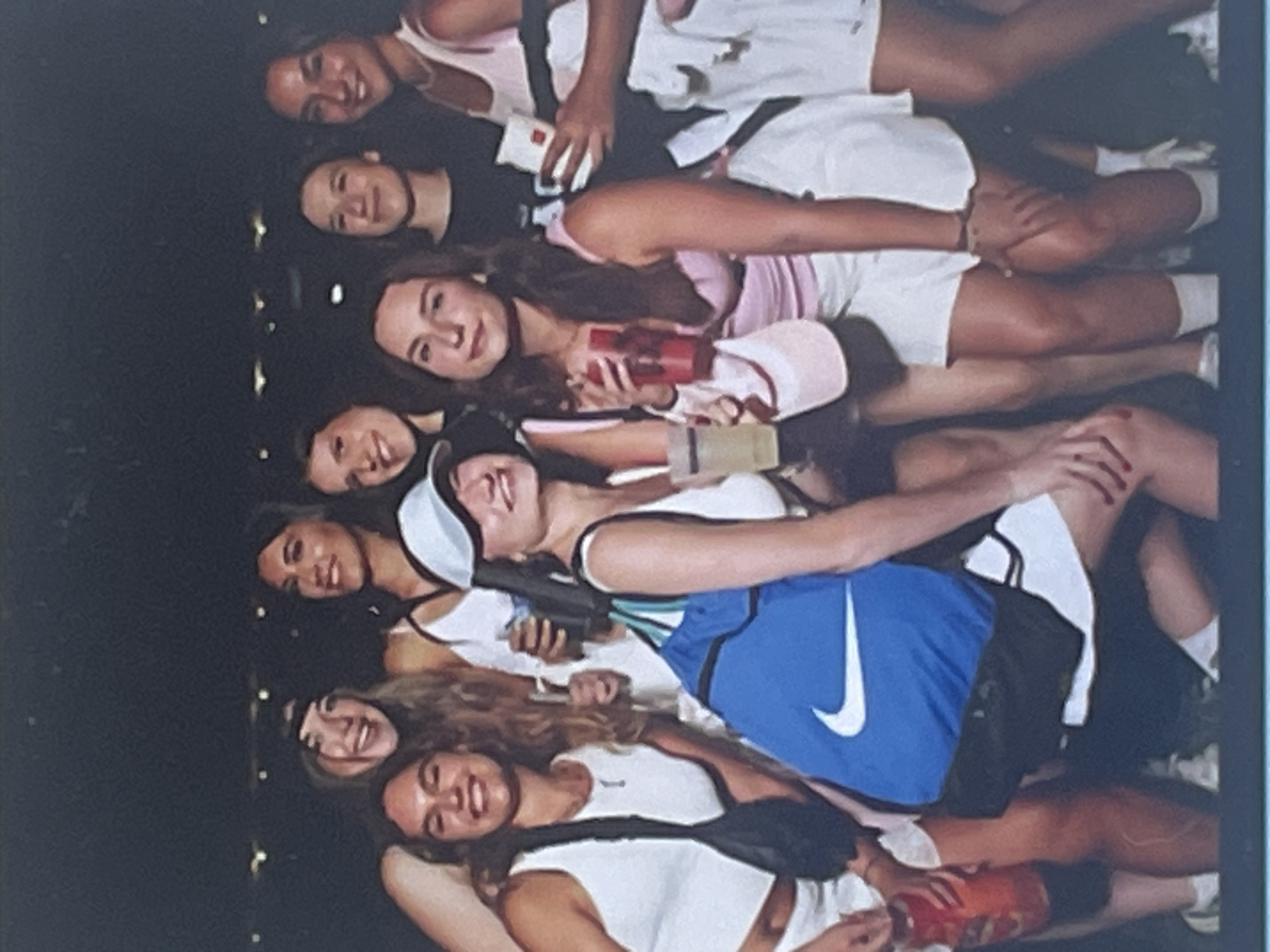 A group of approximately ten young women are arranged closely together in a dimly lit, indoor setting, likely an evening social gathering or party. The subjects are mostly smiling and looking towards the camera, conveying a cheerful and friendly atmosphere.
They are dressed in casual attire, featuring light-colored shorts and tops, with some individuals wearing pink or black tops. Notable accessories include a white visor on one woman's head and a black fanny pack worn by another. Several women are holding red plastic cups, and at least one is holding a clear beverage cup. Mobile phones are also visible in some hands.
A prominent feature is a blue top with a white 'Nike' swoosh logo worn by one woman, and a matching blue drawstring bag with the same 'Nike' logo is seen next to her. The background is dark and out of focus, with blurred light sources suggesting atmospheric lighting typical of an event venue or nightclub. No specific location cues are identifiable within the image itself.