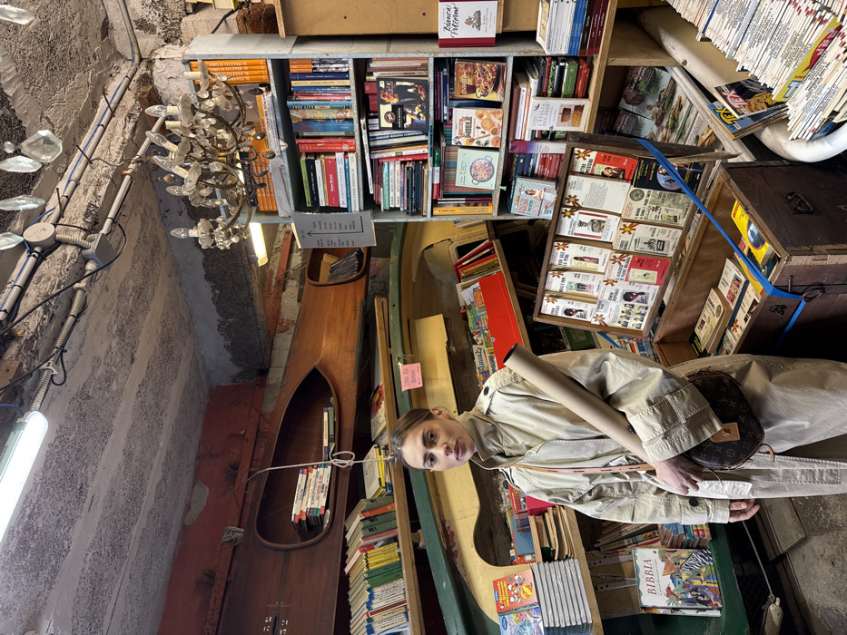 The image depicts a female subject standing within Libreria Acqua Alta, a bookstore located in Venice, Italy. The woman, positioned slightly left of center, faces the viewer directly. She is attired in a high-collared, light beige trench coat, carries a rolled brown paper tube in her left arm, and has a dark brown monogrammed crossbody bag with a light-colored strap slung over her right shoulder. Her hair is light brown and pulled back. The bookstore environment is characterized by densely packed books displayed on shelves, in stacks, and uniquely within wooden boats. A large wooden gondola hull is mounted horizontally on the left wall, functioning as a bookshelf. Below it, a smaller green-painted boat hull also holds books. Tall metal bookshelves filled with a variety of books line the right side of the frame. In the lower foreground, an open wooden trunk containing books is visible, topped by a freestanding display rack holding numerous small publications. A portion of a white bathtub, used for storing books, is discernible on the far right. The ceiling is rough concrete, featuring exposed electrical conduits, a fluorescent tube light, and two suspended crystal chandeliers. Several small informational signs are present throughout the displays.