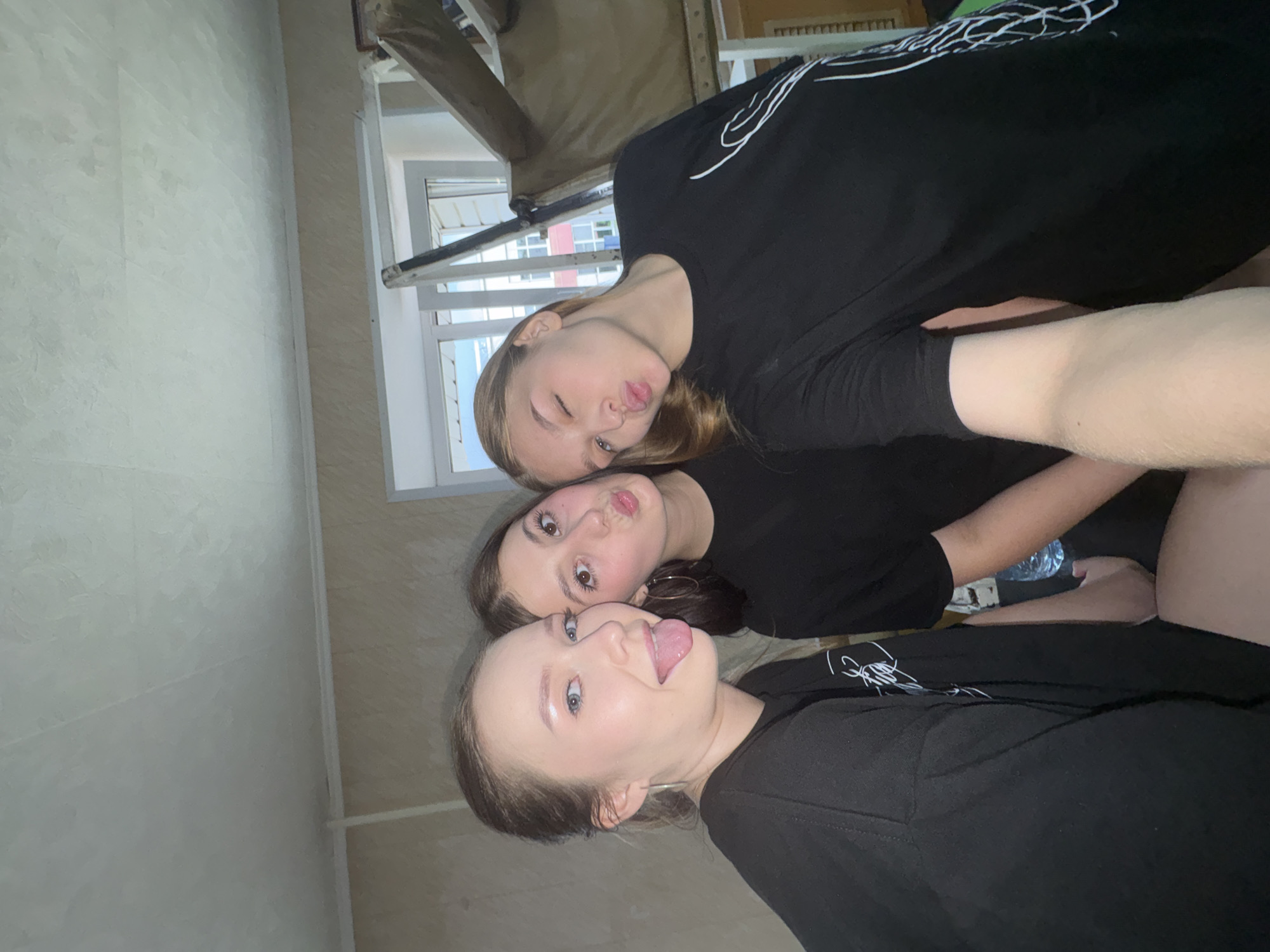Three young women, appearing to be in their late teens or early twenties, are posing for a selfie in an indoor setting. The woman on the left has blonde hair pulled back and is sticking her tongue out, wearing a black long-sleeved shirt. The woman in the middle has dark brown hair and is puckering her lips, also wearing a black long-sleeved shirt. The woman on the right has blonde hair and is winking while puckering her lips, wearing a black long-sleeved shirt with a white graphic on it. The background suggests a residential interior with textured walls and a window with blinds. The scene is consistent with an apartment in Chaikovsky, Russia.