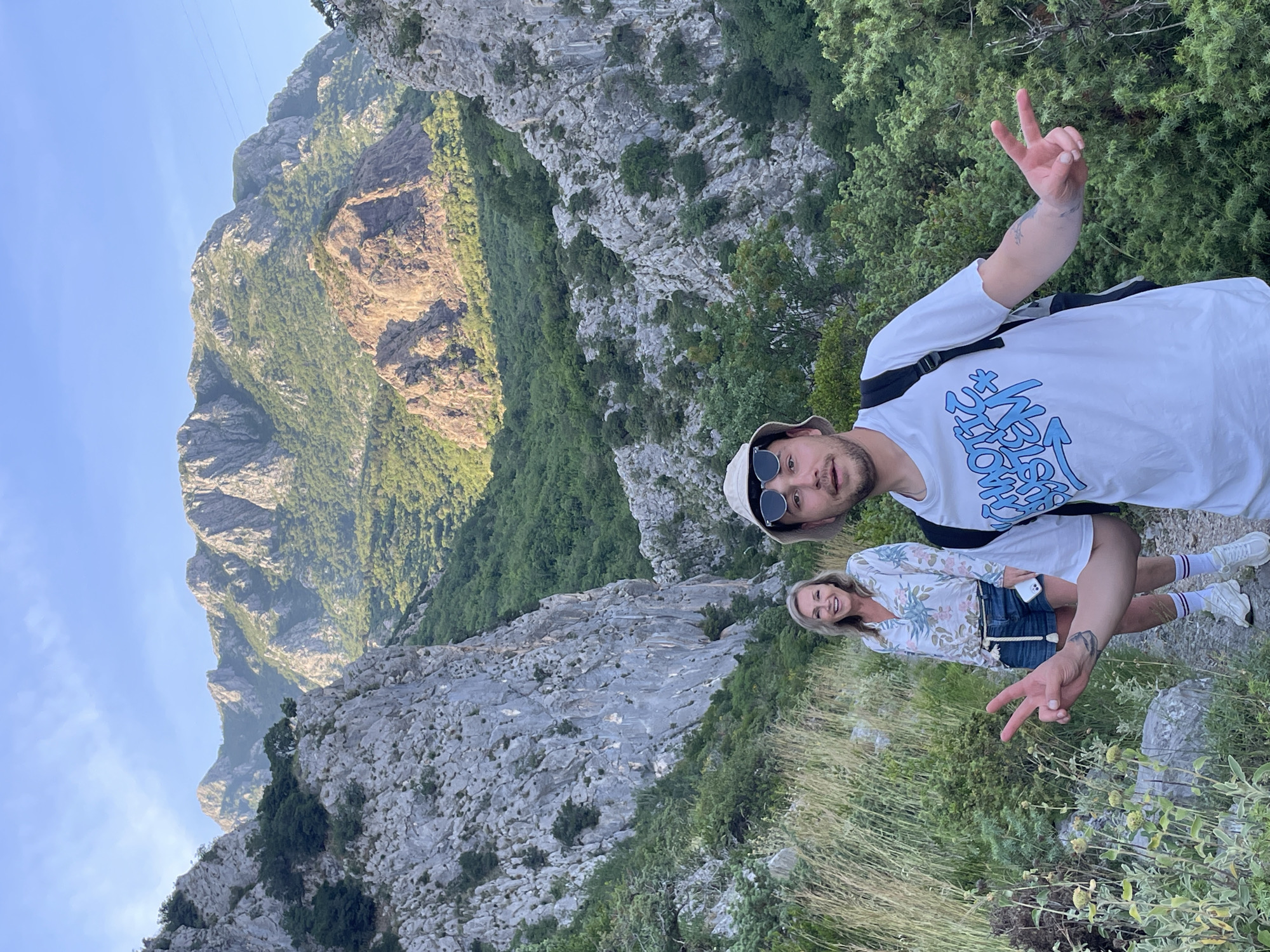 The image, correctly oriented, captures two individuals, a man and a woman, posing on a mountain trail in Zupci, Montenegro, on a clear, sunny day. The background features a striking mountainous landscape with rugged, rocky cliffs and slopes partially covered in dense green vegetation, descending into a valley under a bright blue sky.
In the foreground on the left, a man looks directly at the camera with a relaxed expression. He wears a white short-sleeved t-shirt with stylized blue graphic text, a light-colored bucket hat with sunglasses perched on top, and dark pants. A dark backpack strap is visible over his shoulder, and a tattoo is present on his right forearm. He holds up his right hand, making a peace sign.
To his right and slightly behind him, a woman smiles at the camera, also forming a peace sign with her left hand. She has blonde hair and is dressed in a white long-sleeved blouse with a green and pink floral pattern, denim shorts, white sneakers, and white socks. A mobile phone is tucked into her left shorts pocket.
Both individuals appear joyful and at ease, enjoying the scenic outdoor setting. The ground they stand on is a mix of dry grass and scattered rocks. The mountains showcase varying textures of exposed rock faces and areas densely covered with trees and bushes, suggesting a natural and wild environment. The visible text on the man's shirt is stylized in blue, making specific words difficult to discern.