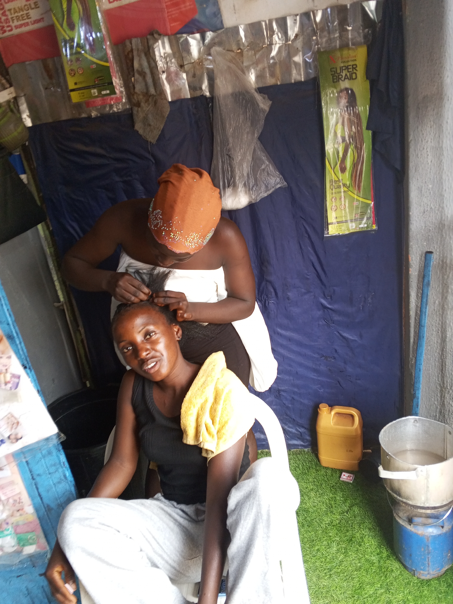 In Gwong, Nigeria, an indoor scene shows two African women. One woman, wearing a brown head covering adorned with shiny embellishments and a white towel draped over her shoulders, stands behind a seated woman and is braiding her hair. The seated woman is wearing a black tank top and gray pants and has a yellow towel draped over one shoulder.
The setting includes a blue tarp backdrop and several containers, including a metal bucket on a blue and silver stove. Hair product packaging hangs on the tarp. Artificial green turf covers the floor area. The location appears to be a modest, improvised hair salon.