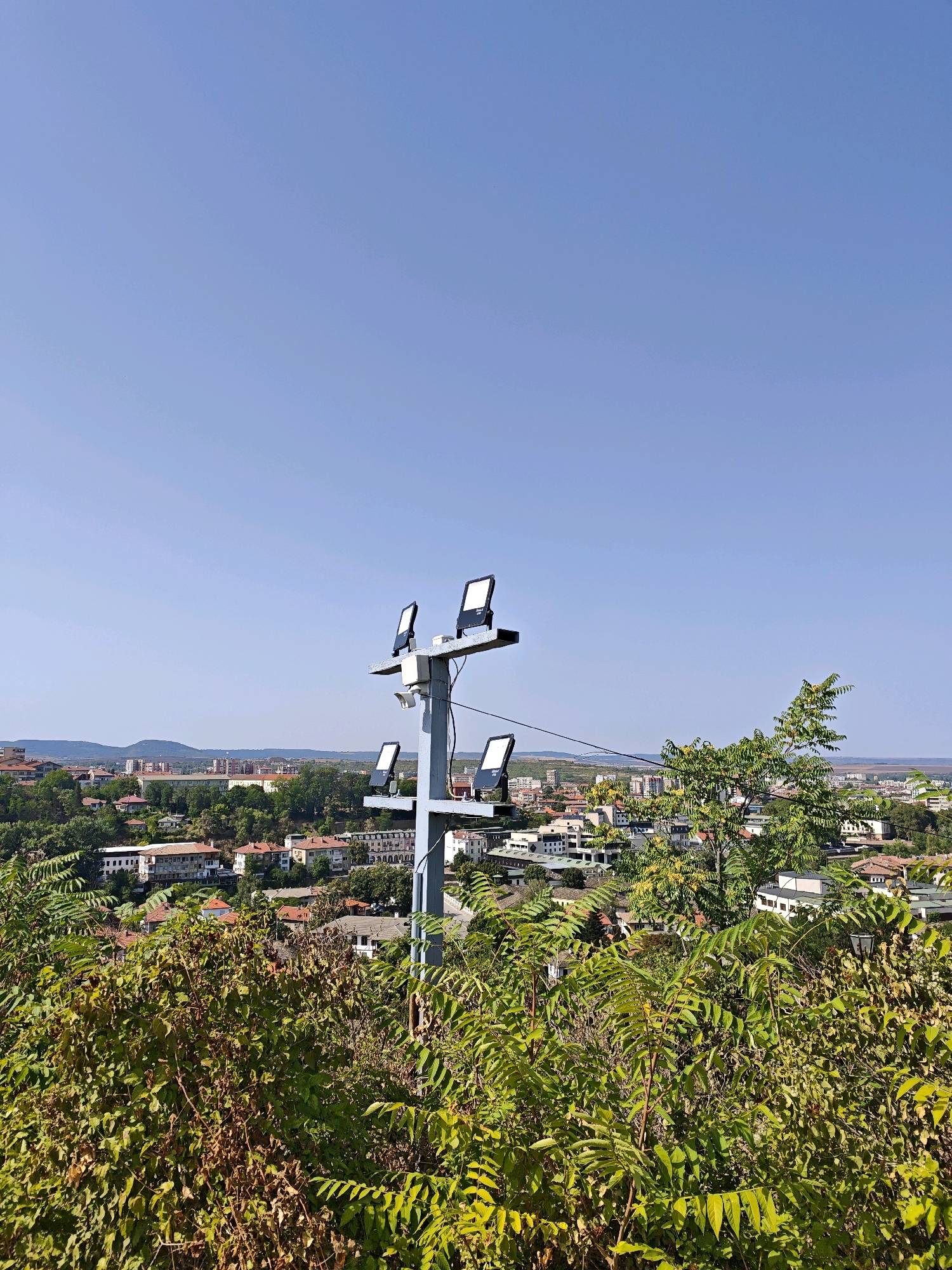 The image displays an elevated view of the city of Lovech, Bulgaria, on a clear, sunny day. In the foreground, dense green foliage partially obscures the view. A prominent utility pole, painted gray, stands near the center of the frame, topped with several modern, rectangular floodlights. A small white surveillance camera is mounted on the pole below the lights. Behind the foreground vegetation, the city unfolds with a mix of buildings featuring red-tiled roofs, interspersed with trees. In the distance, rolling hills are visible under a vast, pale blue sky. The scene appears to be a quiet, daytime cityscape, possibly from a park or a higher vantage point overlooking the residential areas.
