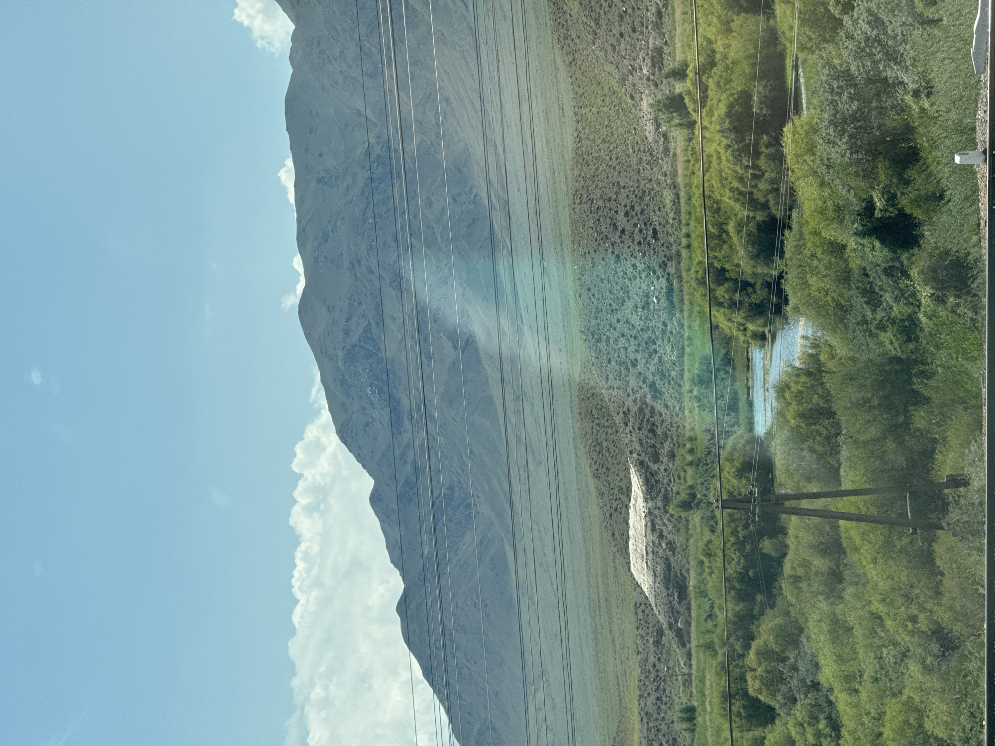 A panoramic, eye-level shot depicts a rural landscape dominated by mountains, vegetation, and a river, located in the city of Kok-Moinok - 1, Kyrgyzstan. The sky is a clear blue with a few scattered white clouds. A range of hazy, muted green mountains stretches across the horizon, their slopes etched with subtle lines and shadows. In the foreground, a lush green valley is filled with dense foliage and trees. A meandering river, reflecting the blue sky, winds through the vegetation. Several power lines are strung across the frame, creating a distinct linear pattern in the middle ground. A single, dark wooden utility pole stands prominently in the lower left quadrant, supporting the power lines. A large, light-colored, tarp-covered mound is visible on a hillside in the mid-ground. The overall impression is one of natural beauty and agricultural or industrial activity.
